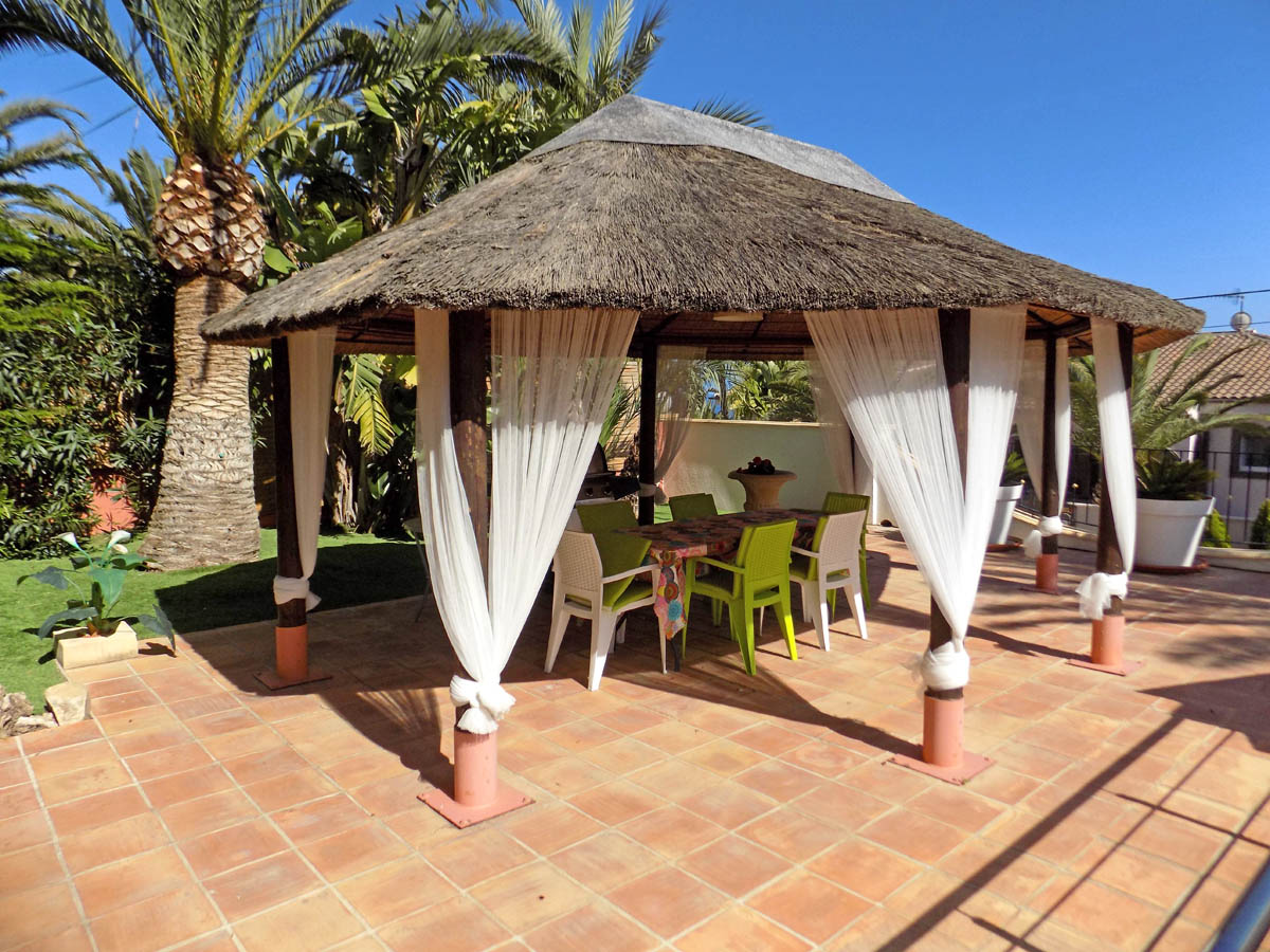 Outdoor gazebo with a thatched roof and decorative white curtains on a terracotta terrace, featuring a dining area amidst Mediterranean palms.