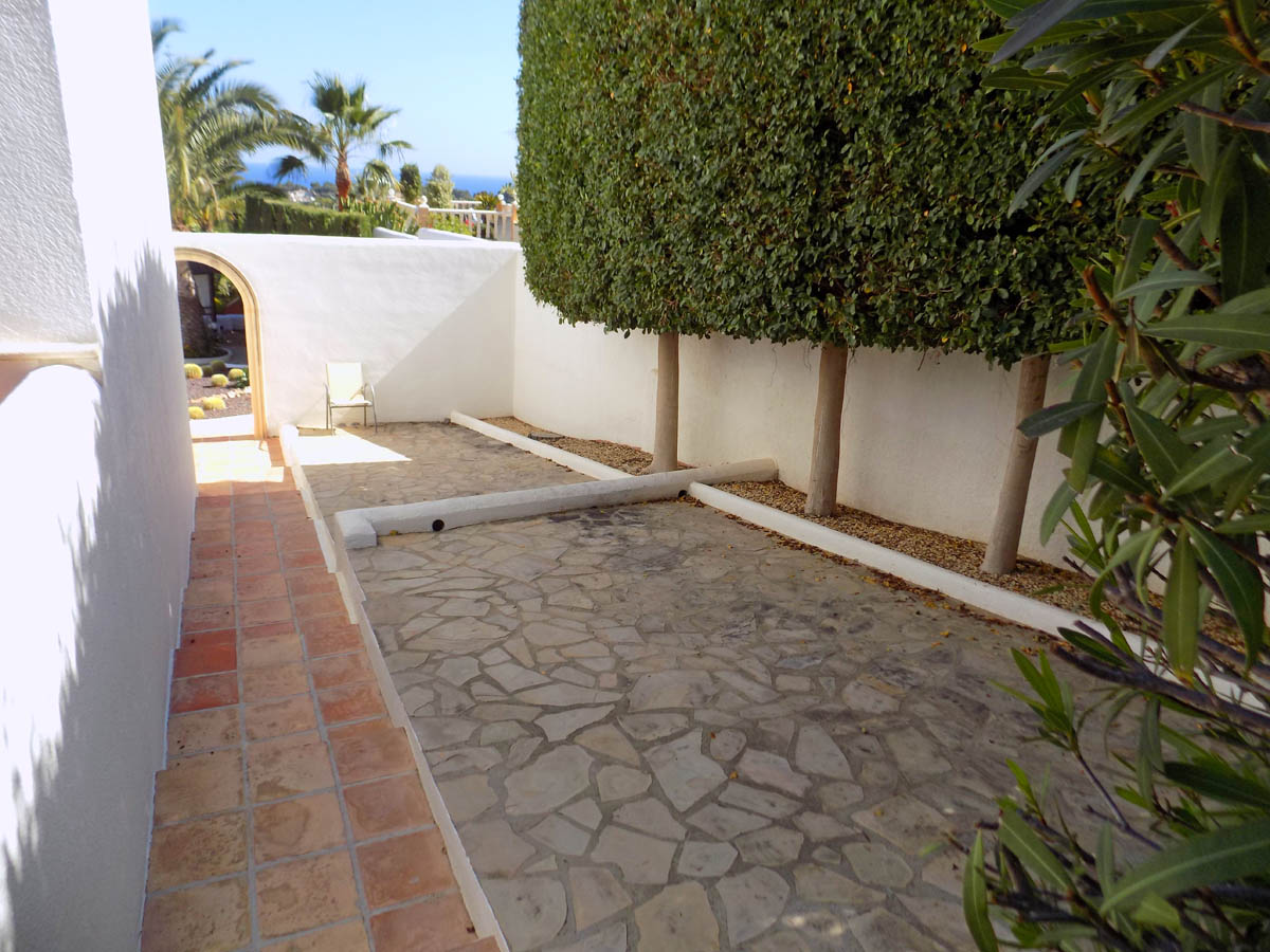 Outdoor patio with natural stone paving and white walls, bordered by high manicured hedges. A terracotta tiled walkway leads towards an arched garden entrance.