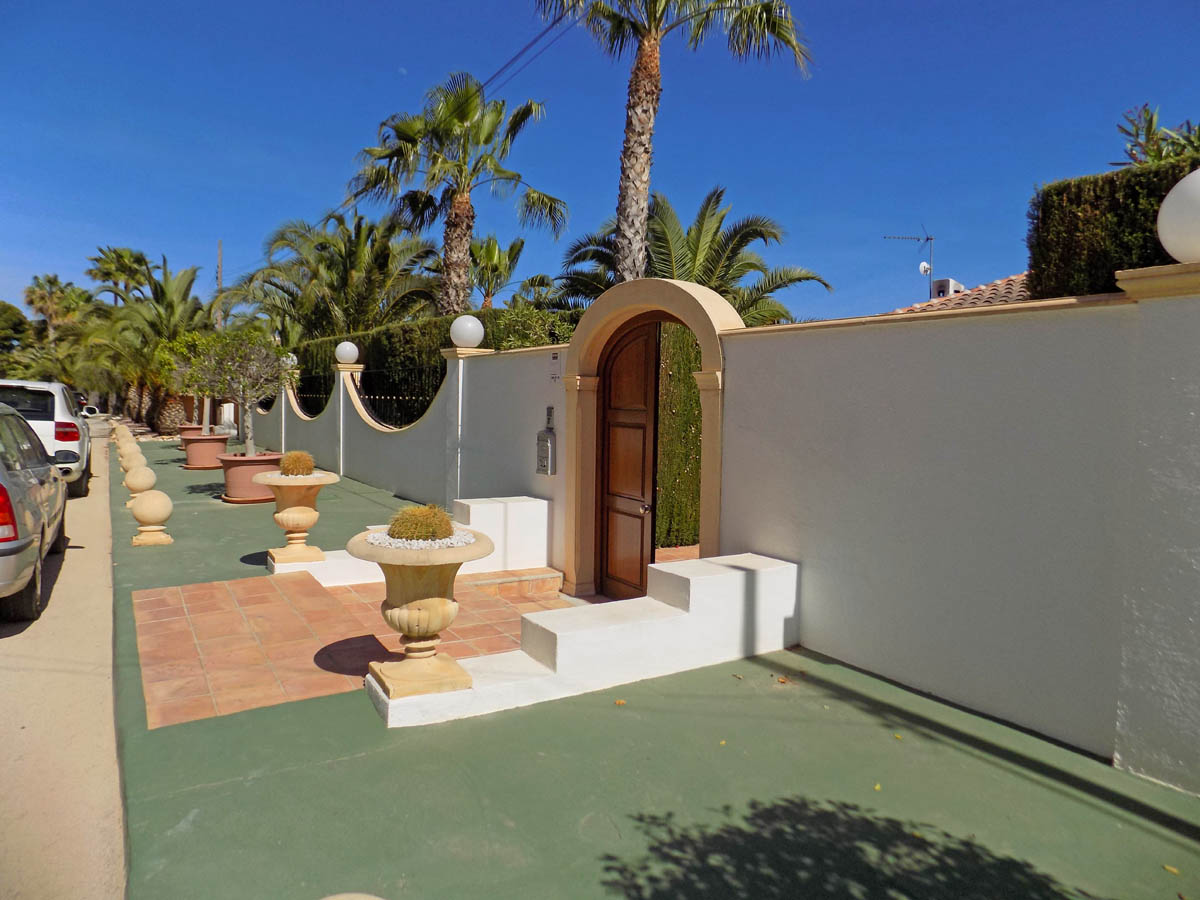 Pedestrian entrance featuring an arched wooden door, white perimeter wall with decorative stone planters, and lush palm trees.