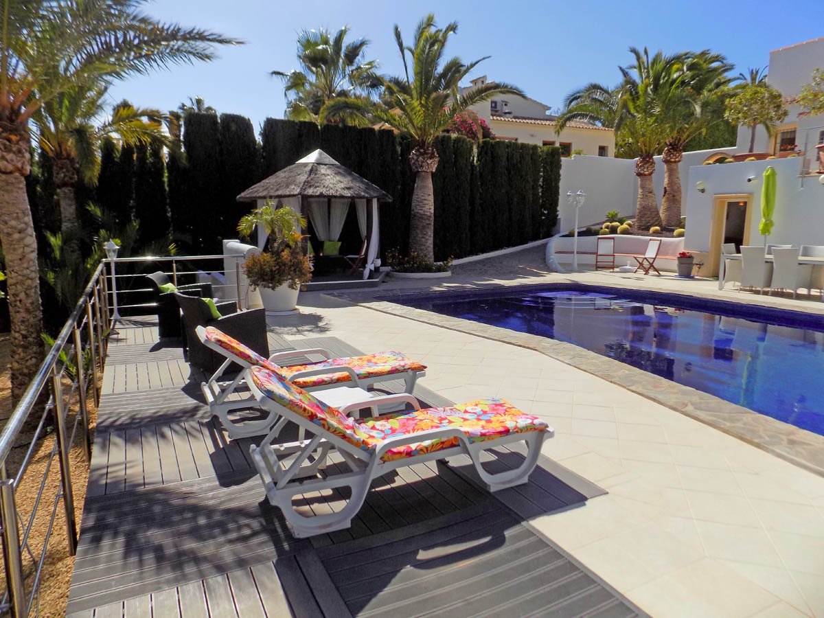 Pool area featuring a combination of stone and composite decking, sun loungers, a thatched gazebo with curtains, and mature palm trees.