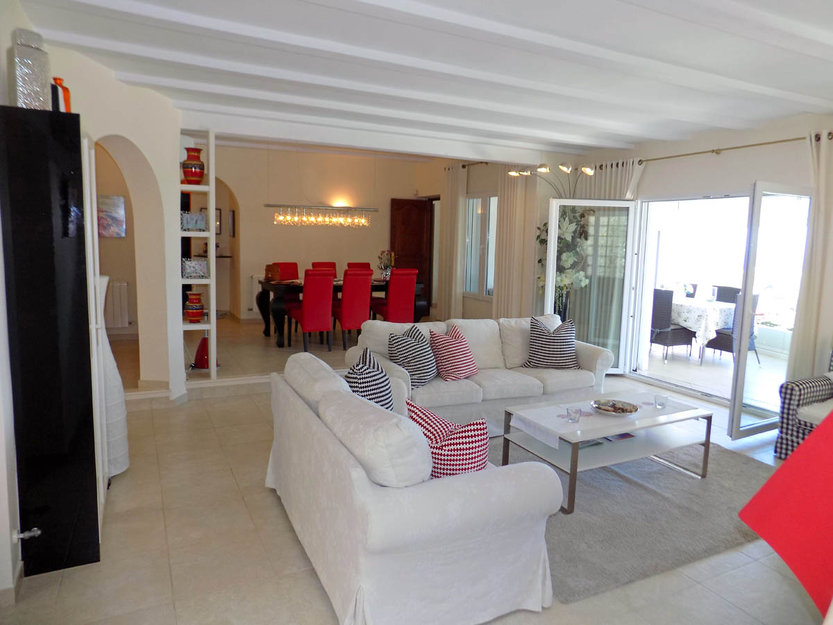 Spacious living area featuring white beamed ceilings, neutral sofas, and a dining zone with vibrant red chairs. Large glass doors open onto the bright glazed terrace.