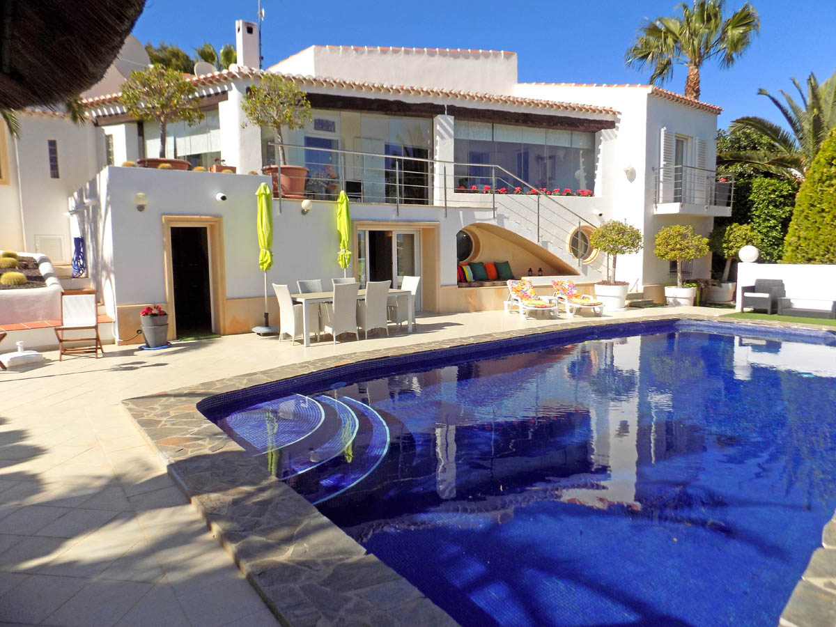 Villa exterior featuring a large blue mosaic pool, stone-paved sun terrace with outdoor dining area, and a glazed upper porch overlooking the Mediterranean garden.
