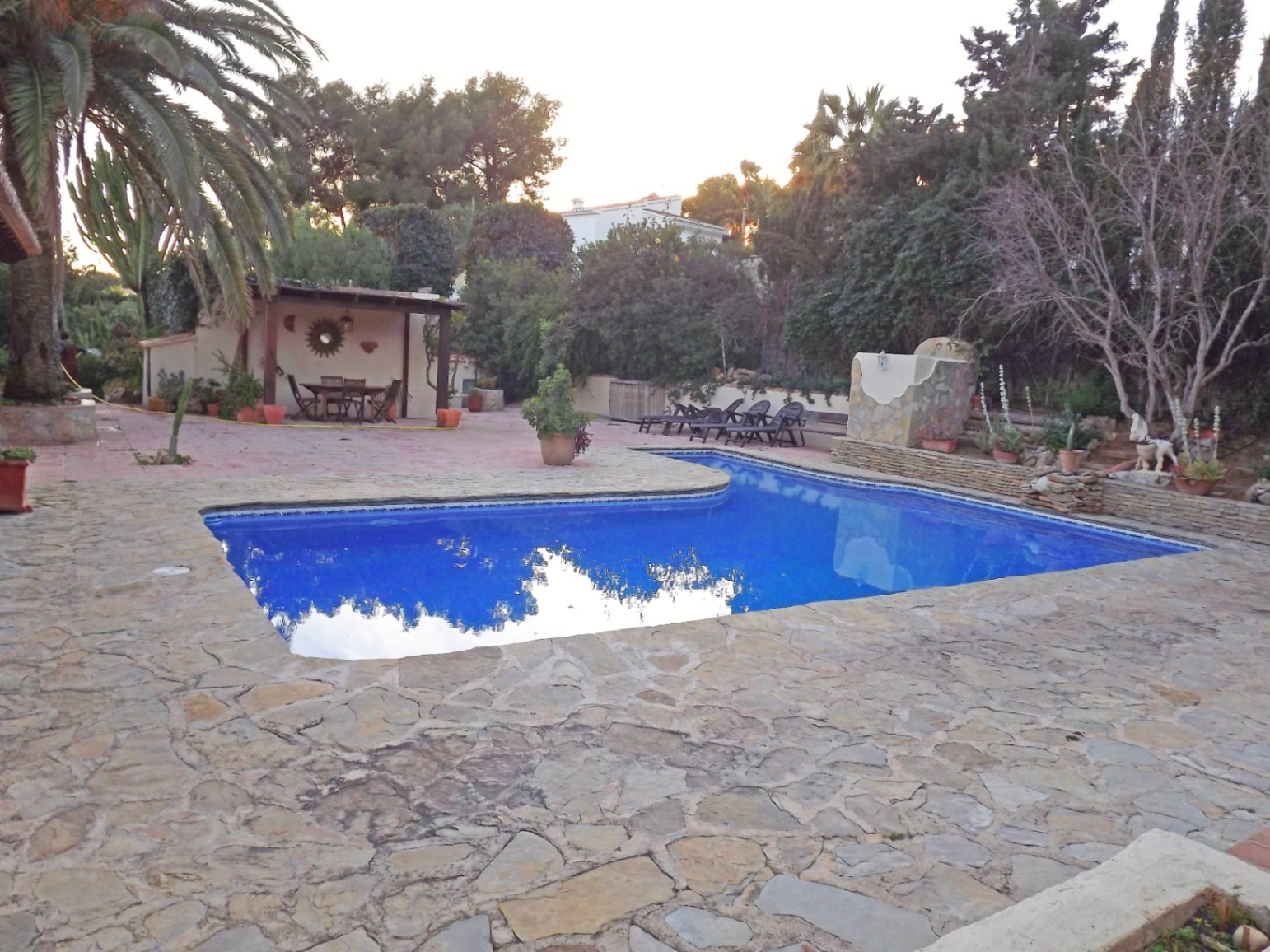 A 12x5m swimming pool featuring a natural stone terrace, a covered outdoor dining area with wooden beams, and lush Mediterranean landscaping.