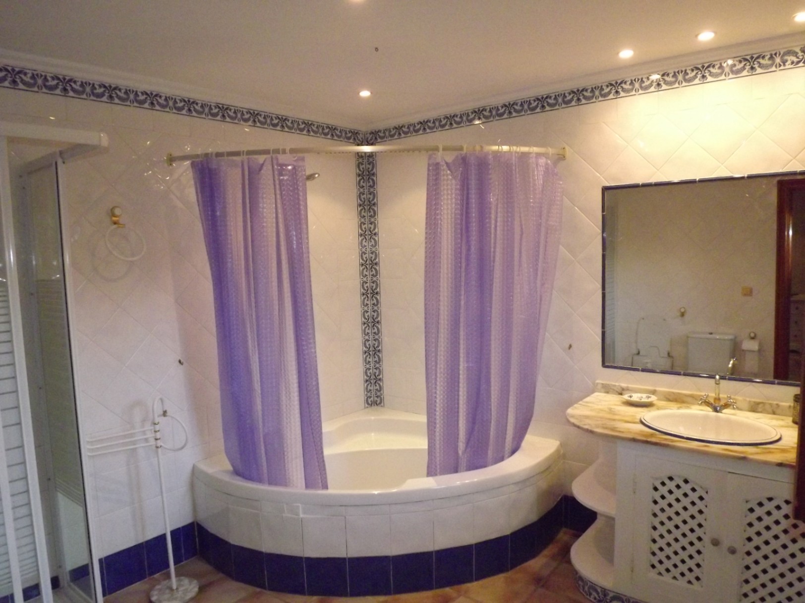 Bathroom featuring a corner bathtub with purple curtains and white tiling with blue decorative borders. A marble-topped vanity and large mirror complete the space.