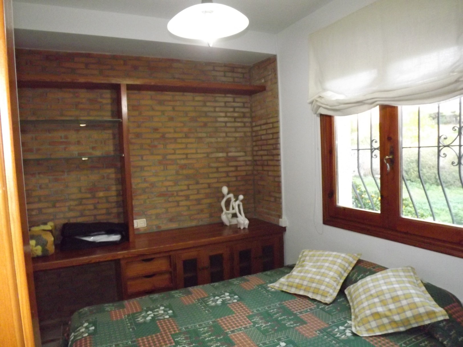 Bedroom featuring an exposed brick accent wall, integrated wooden cabinetry with glass shelving, and a wooden window frame overlooking the garden.