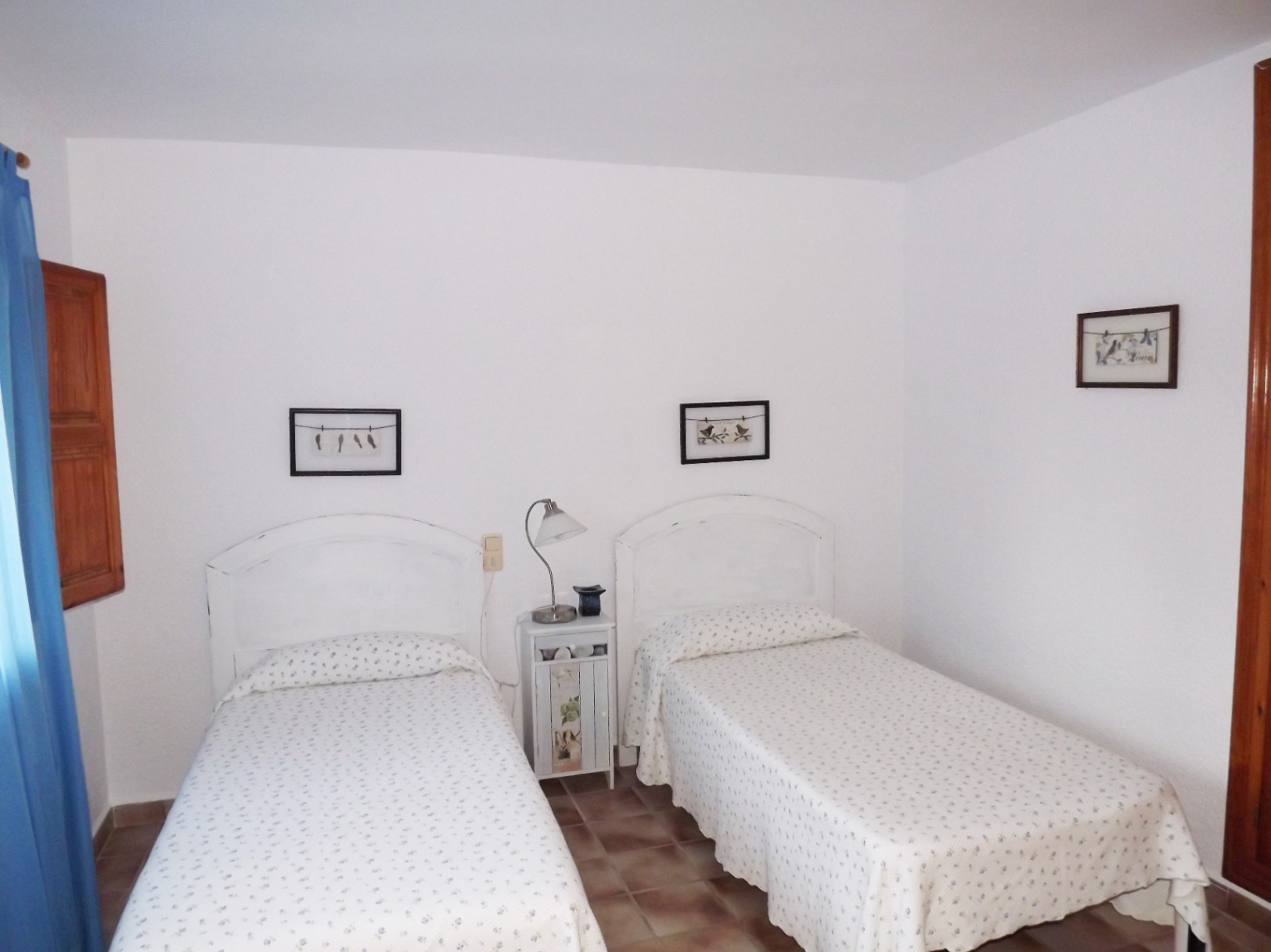 Bedroom featuring twin beds with white arched headboards and floral linens. The space includes terracotta tile flooring, a central nightstand, and a window with wooden shutters.