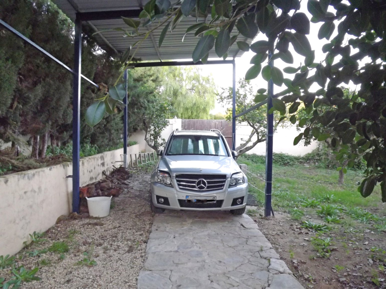Covered parking area featuring a metal carport structure and natural stone paving, set within the property's landscaped garden grounds.