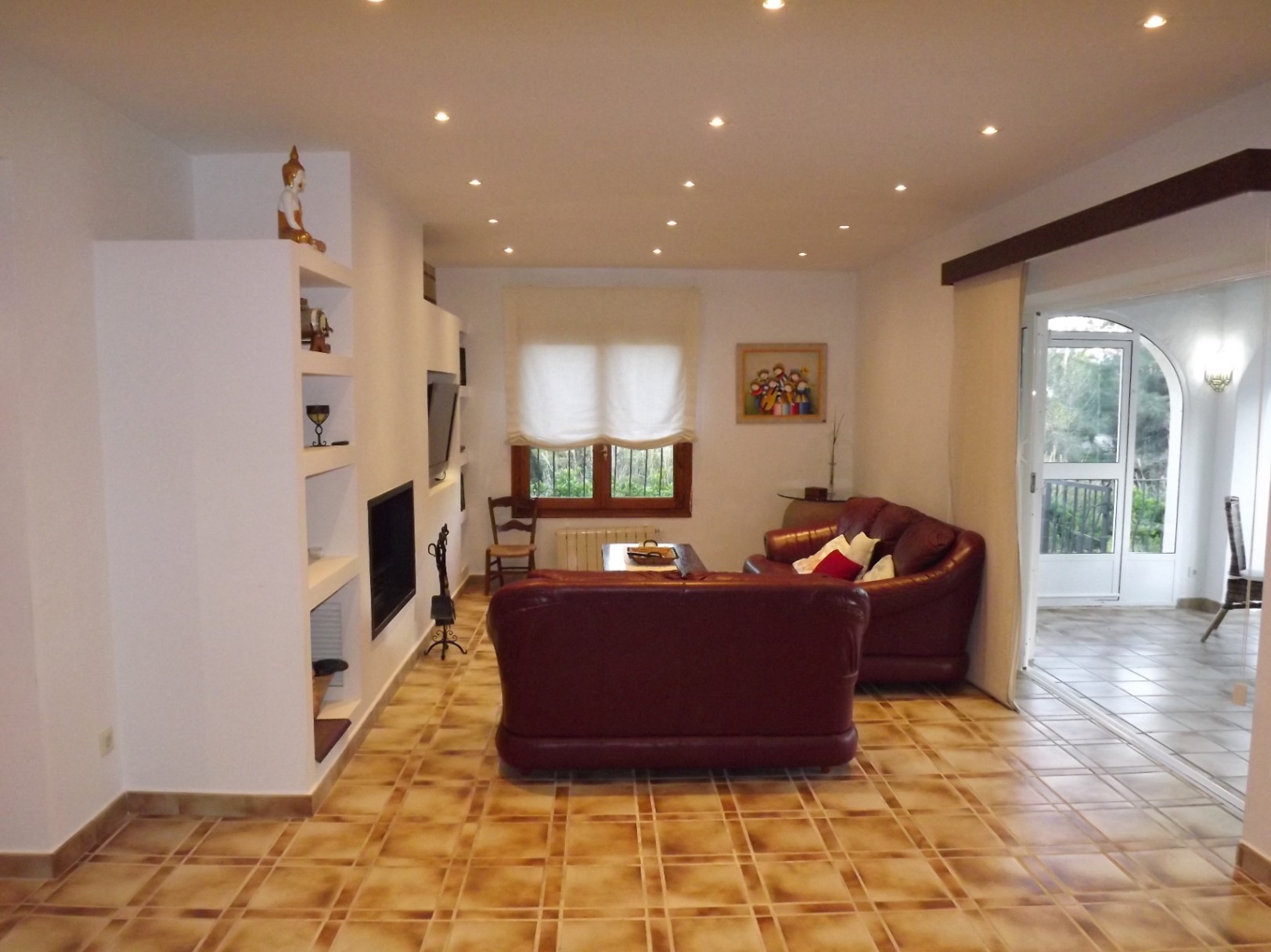 Cozy living room featuring a built-in fireplace, burgundy leather sofas, and tiled flooring. The space includes recessed ceiling lighting and leads to the glazed terrace.