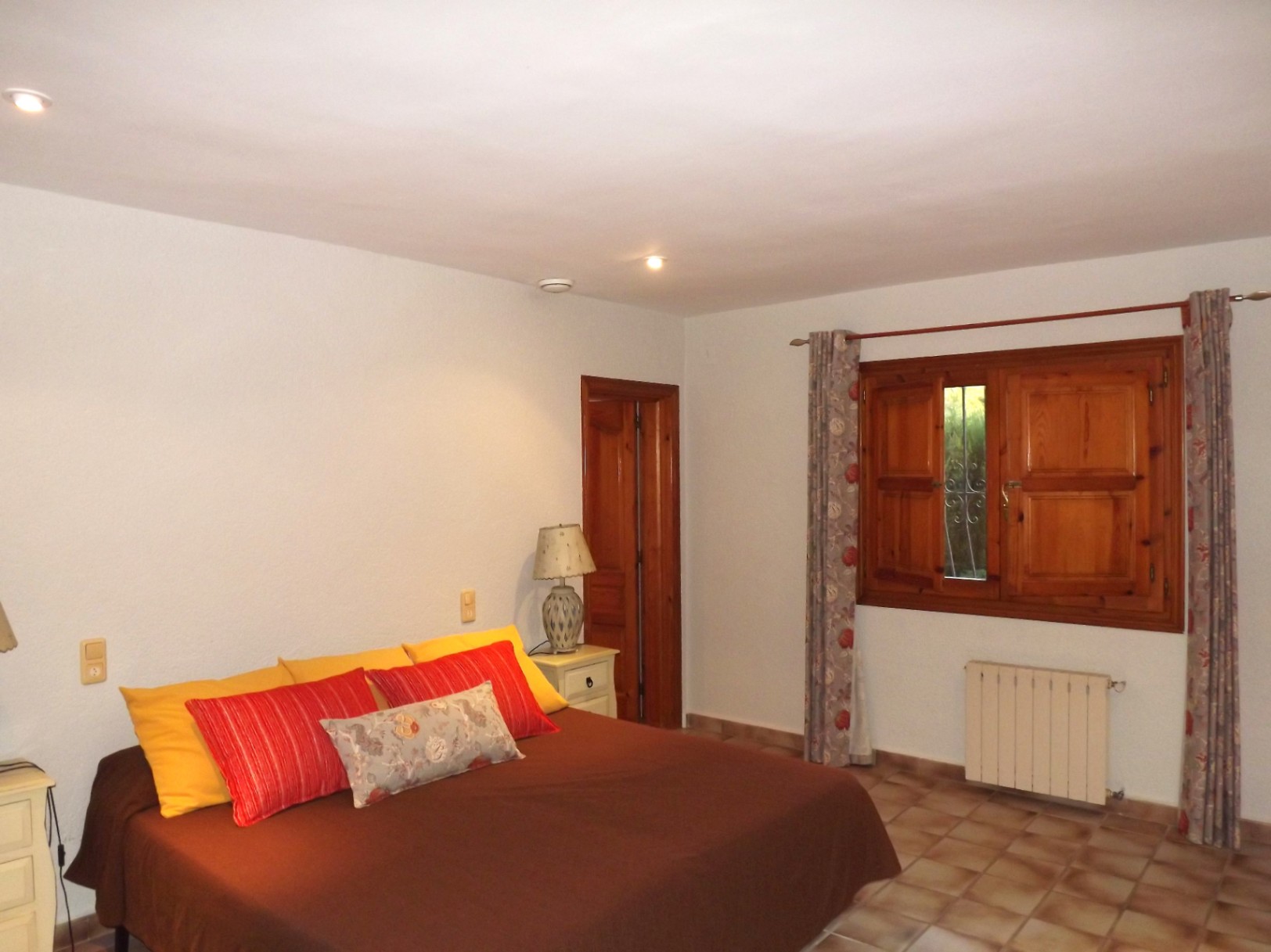 Double bedroom featuring terracotta floor tiles, traditional wooden window shutters, and a central heating radiator. Includes recessed ceiling lighting and solid wood doors.