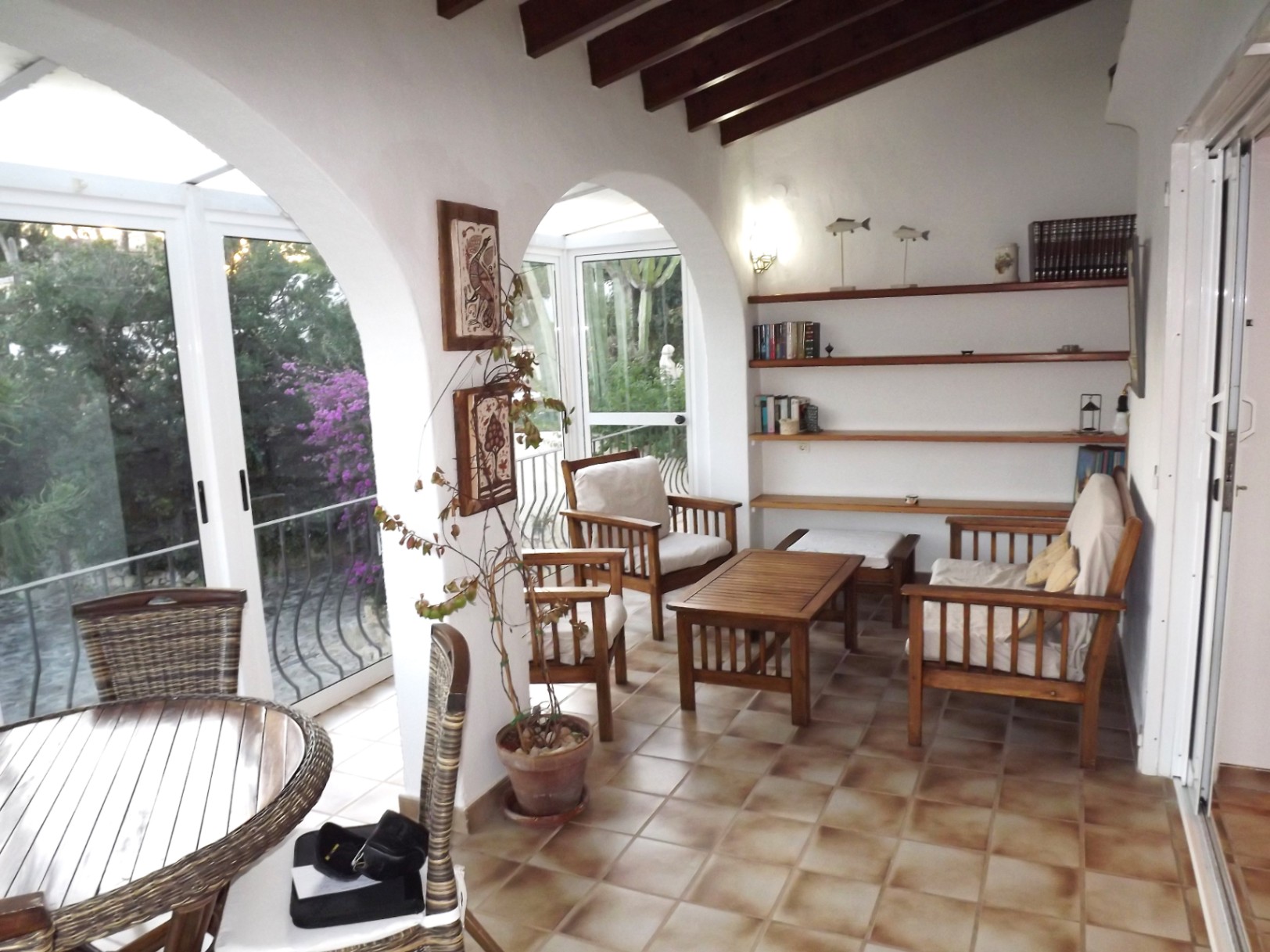 Glazed terrace featuring traditional arches, wooden beam ceilings, and terracotta flooring. Furnished with wooden seating and built-in shelving overlooking the garden.