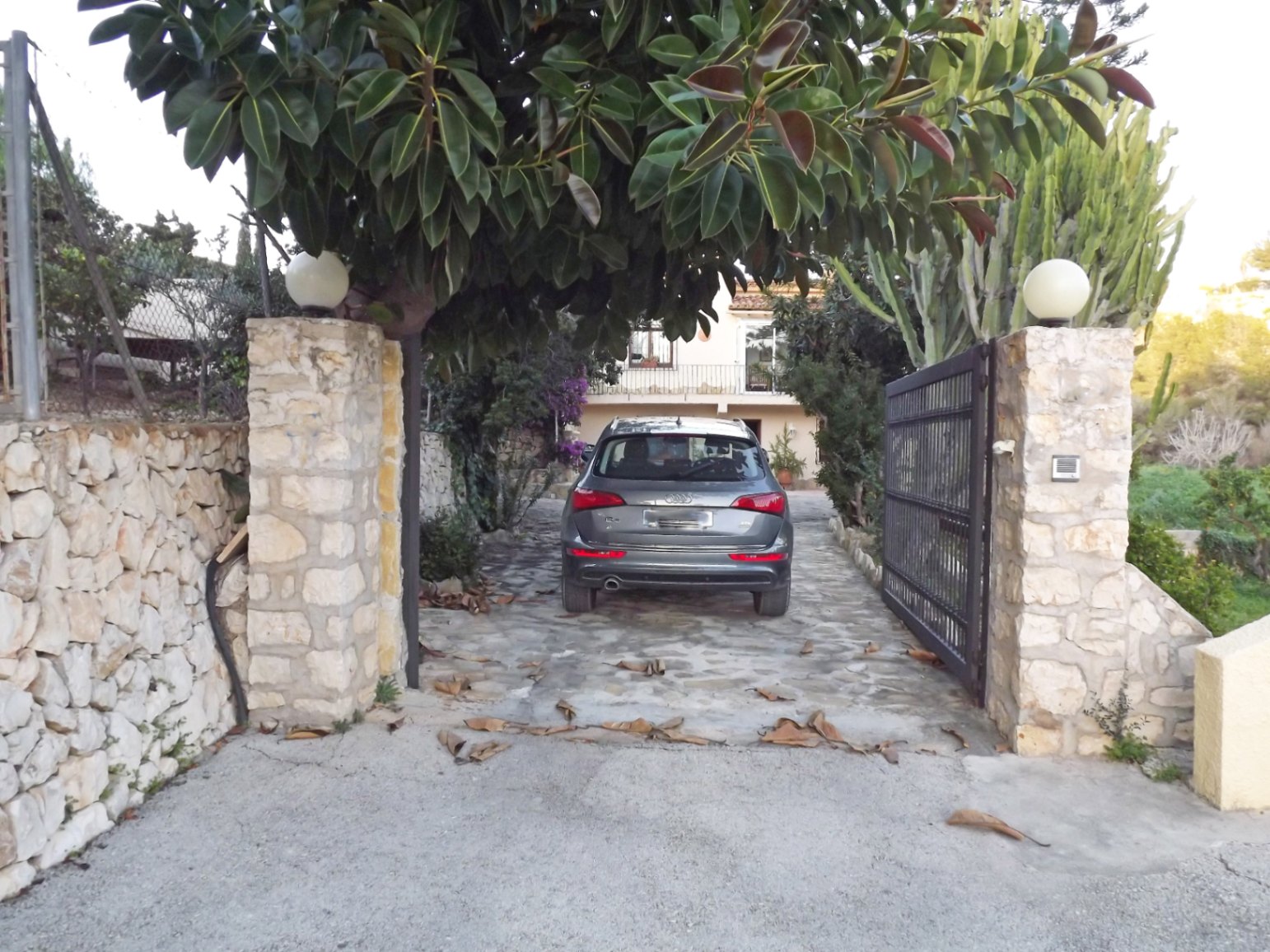 Main entrance featuring natural stone pillars with spherical lamps and a metal gate opening onto a paved driveway shaded by mature Mediterranean trees.