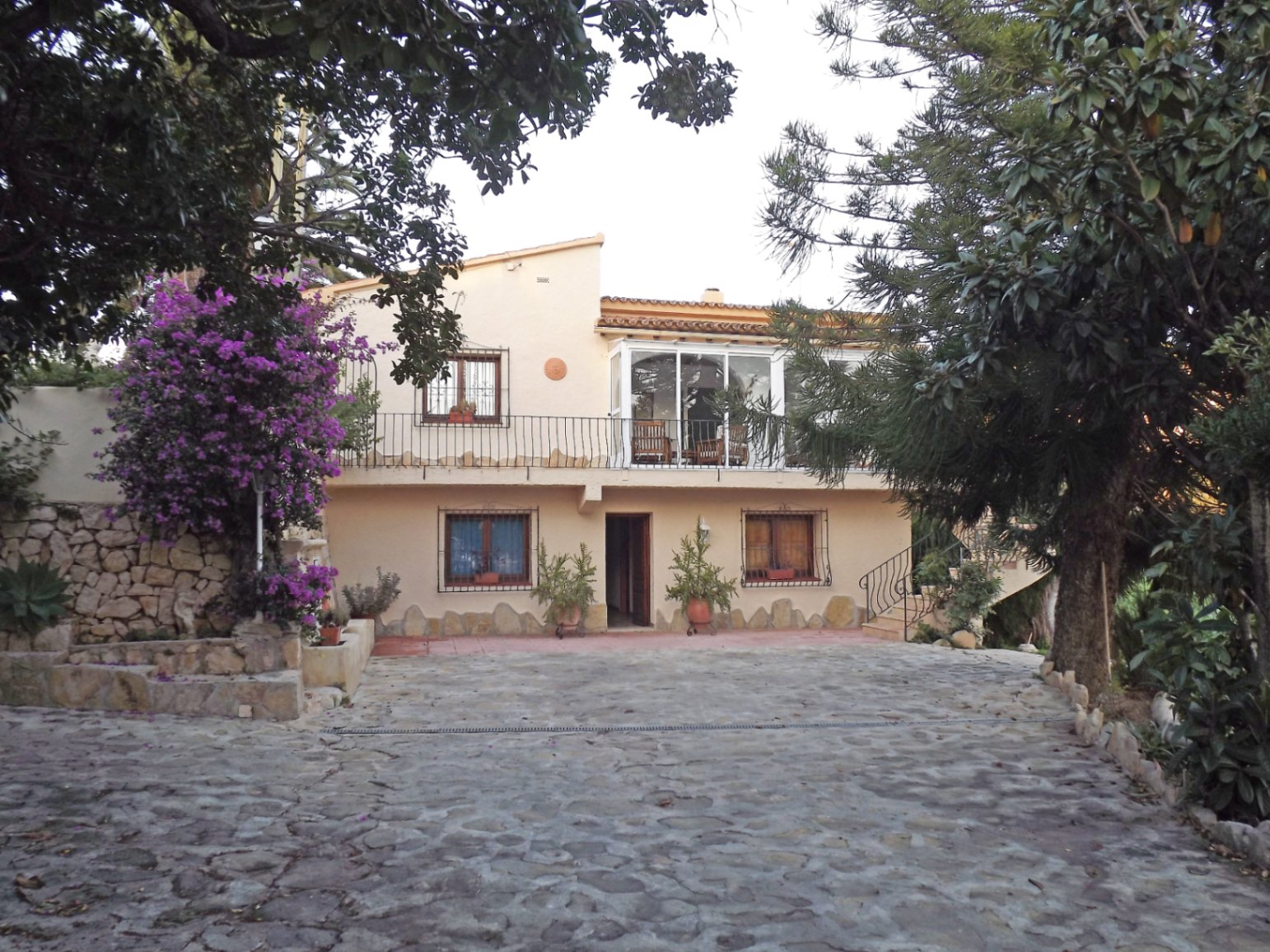 Main facade of the villa featuring a stone-paved driveway, a glazed upper terrace, and traditional Mediterranean architecture with wrought iron details.