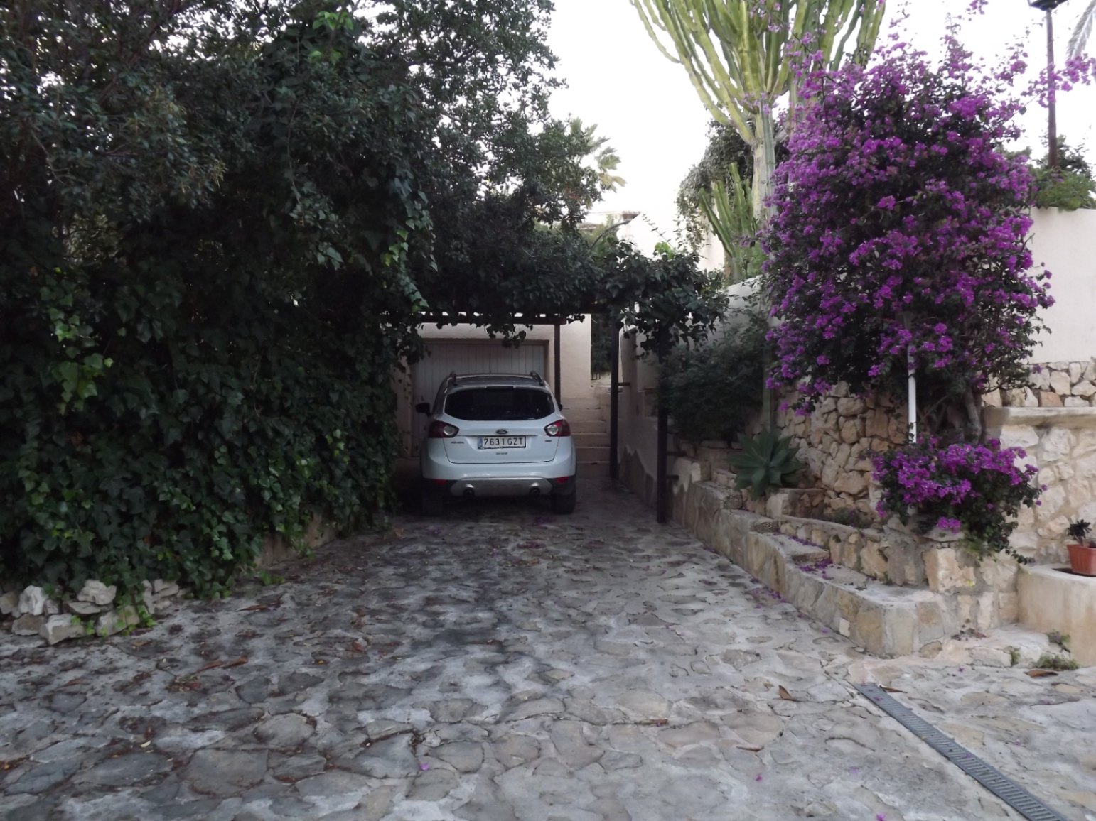 Natural stone driveway leading to the garage, framed by lush Mediterranean greenery, purple bougainvillea, and traditional dry stone walls.