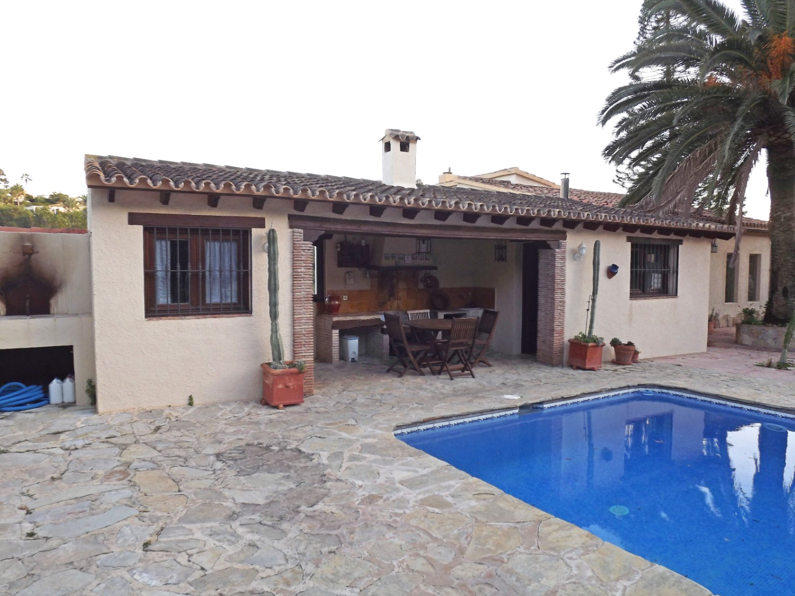 Outdoor terrace featuring natural stone paving, a 12x5m swimming pool, and a covered barbecue area with wooden beams and a dining set next to a palm tree.