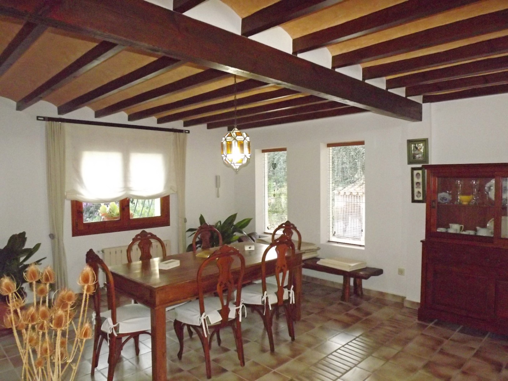 Rustic dining area featuring exposed dark wood ceiling beams, a solid wood table with carved chairs, and traditional terracotta tile flooring.