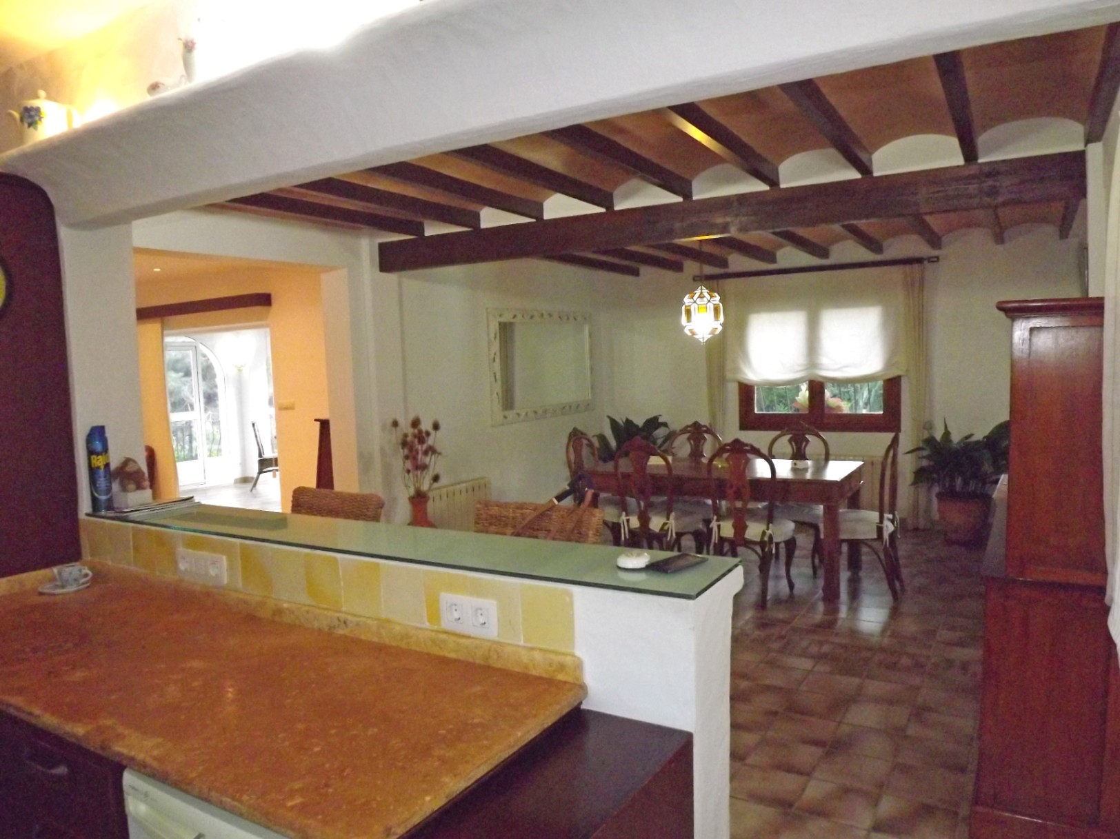 View from the kitchen toward the dining area, featuring exposed dark wooden ceiling beams with terracotta tiles, ceramic flooring, and a stone breakfast bar.