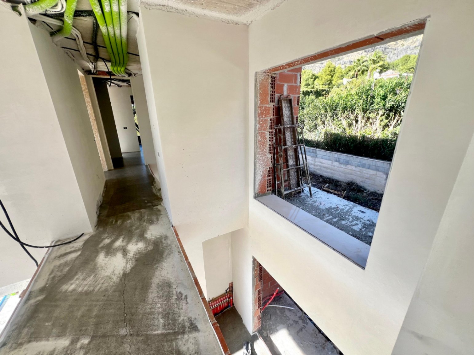 Interior hallway under construction showcasing double-height architecture, exposed technical installations, and a large window opening to the greenery.