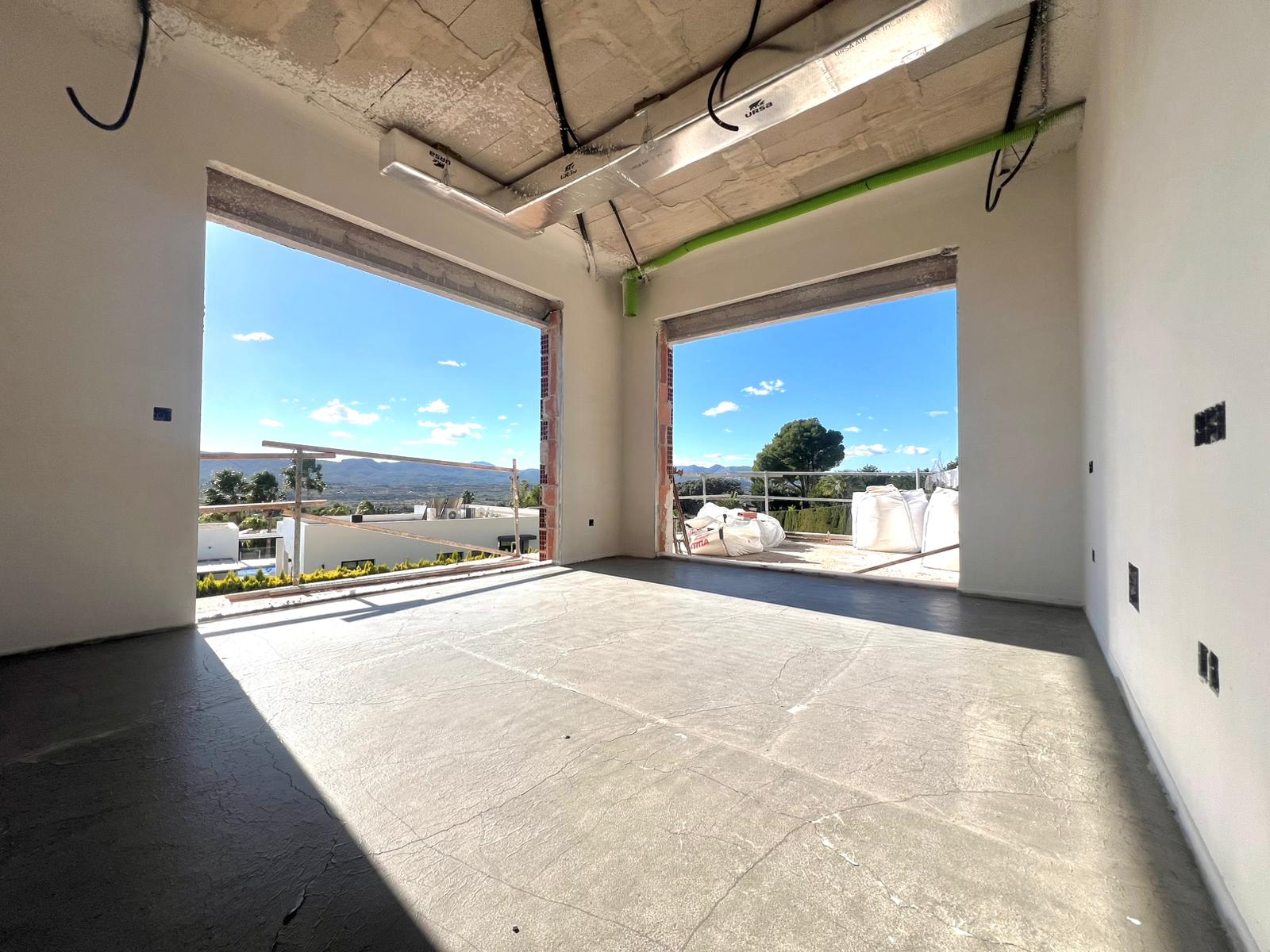 Interior under construction featuring large window openings with panoramic valley views, exposed HVAC ducting, and structural concrete ceilings.