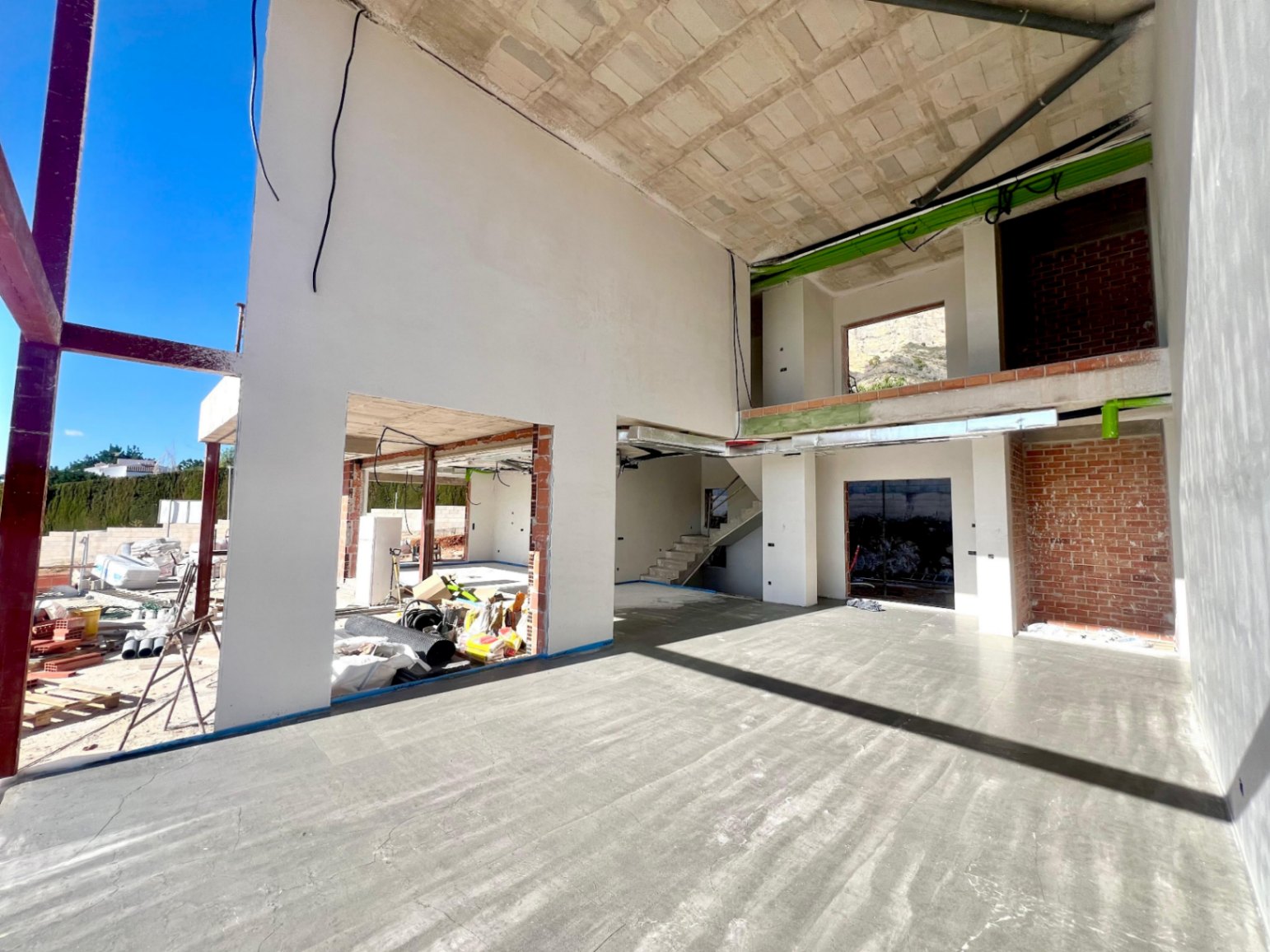Interior view of the double-height living area under construction, featuring the open mezzanine, concrete staircase, and large openings for floor-to-ceiling windows.