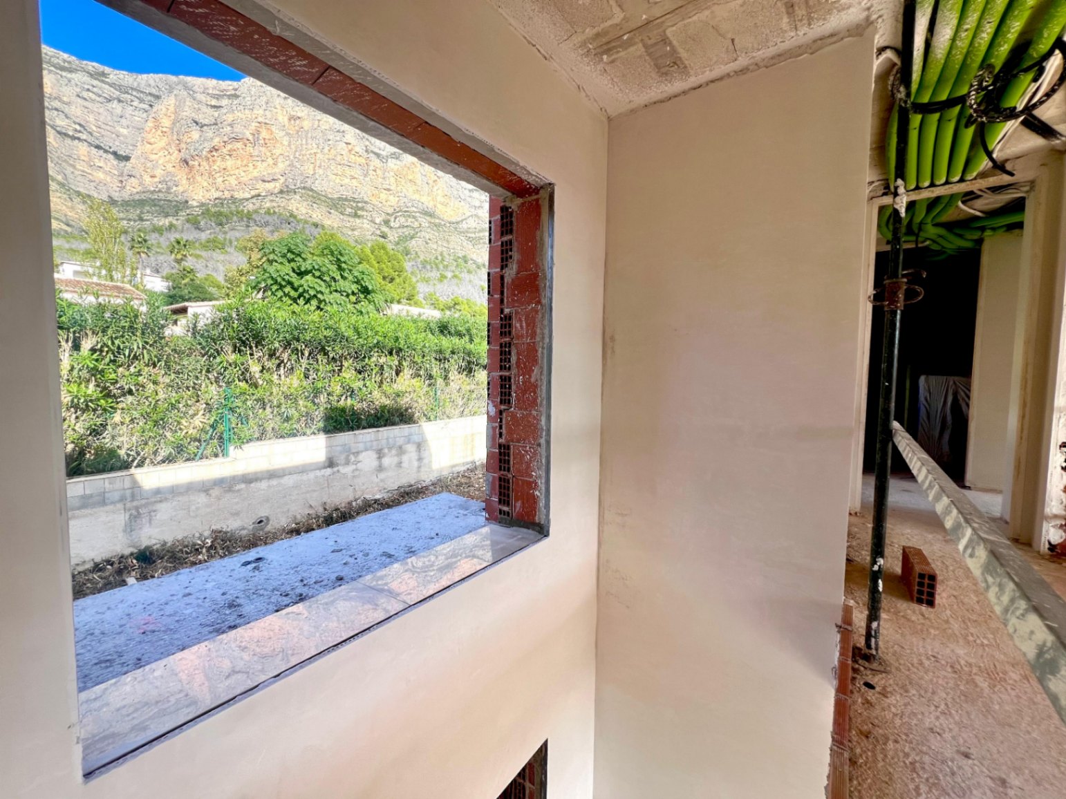Interior view under construction featuring a large window frame overlooking the Montgó mountain, with visible ceiling conduits and plastered walls.