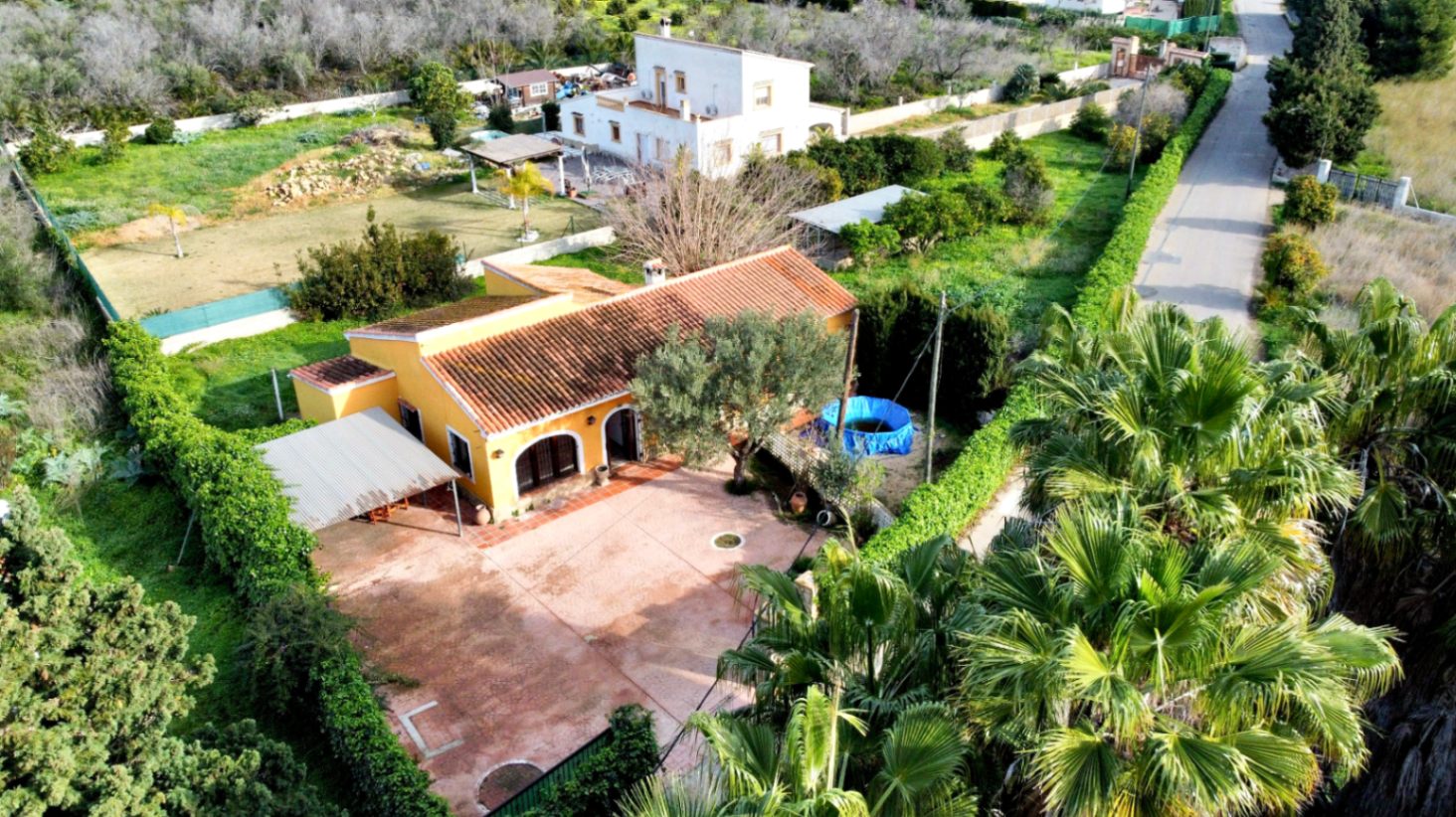 Aerial view of the property featuring the yellow facade, large paved patio, arched porch, and covered parking area surrounded by lush Mediterranean greenery.