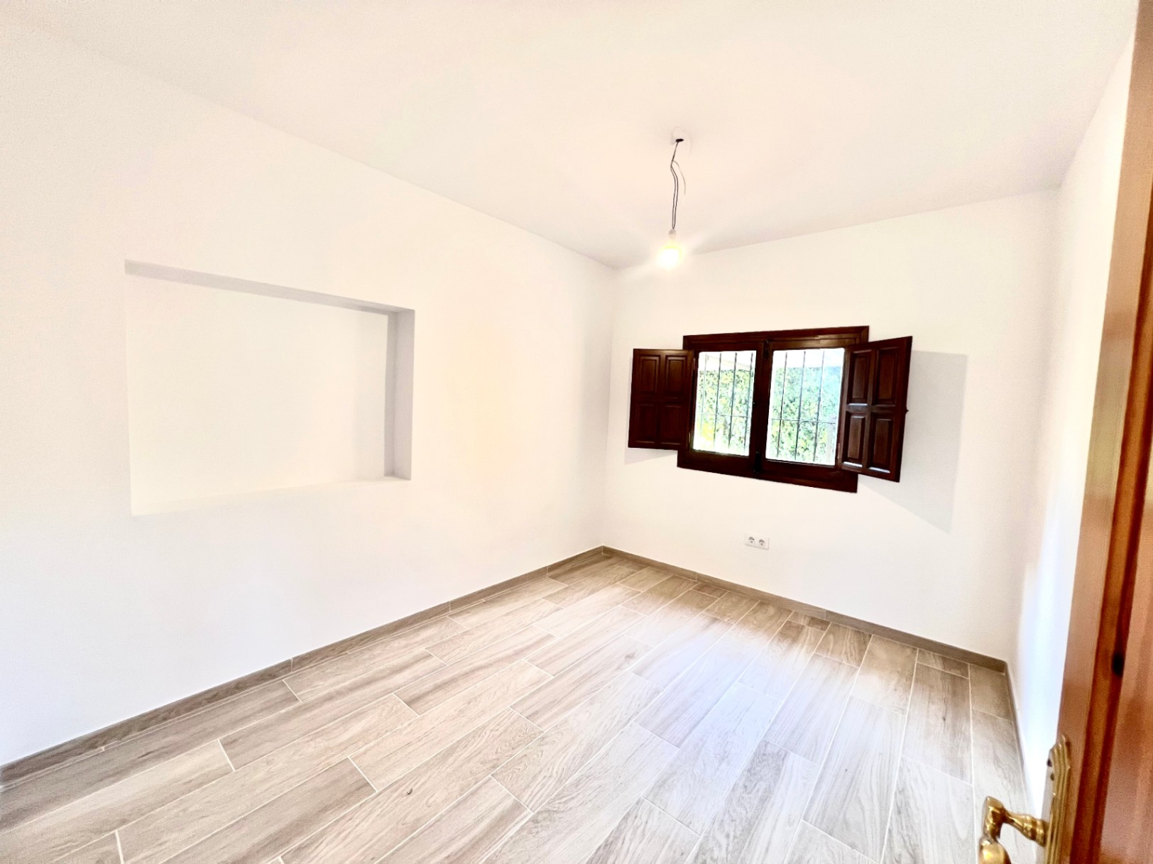 Bright bedroom featuring wood-effect ceramic flooring, white walls, a built-in wall niche, and a window with traditional dark wood shutters.