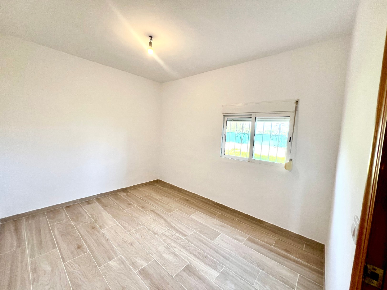 Bright bedroom featuring wood-effect ceramic tile flooring, smooth white walls, and a window equipped with an ornamental security grate.