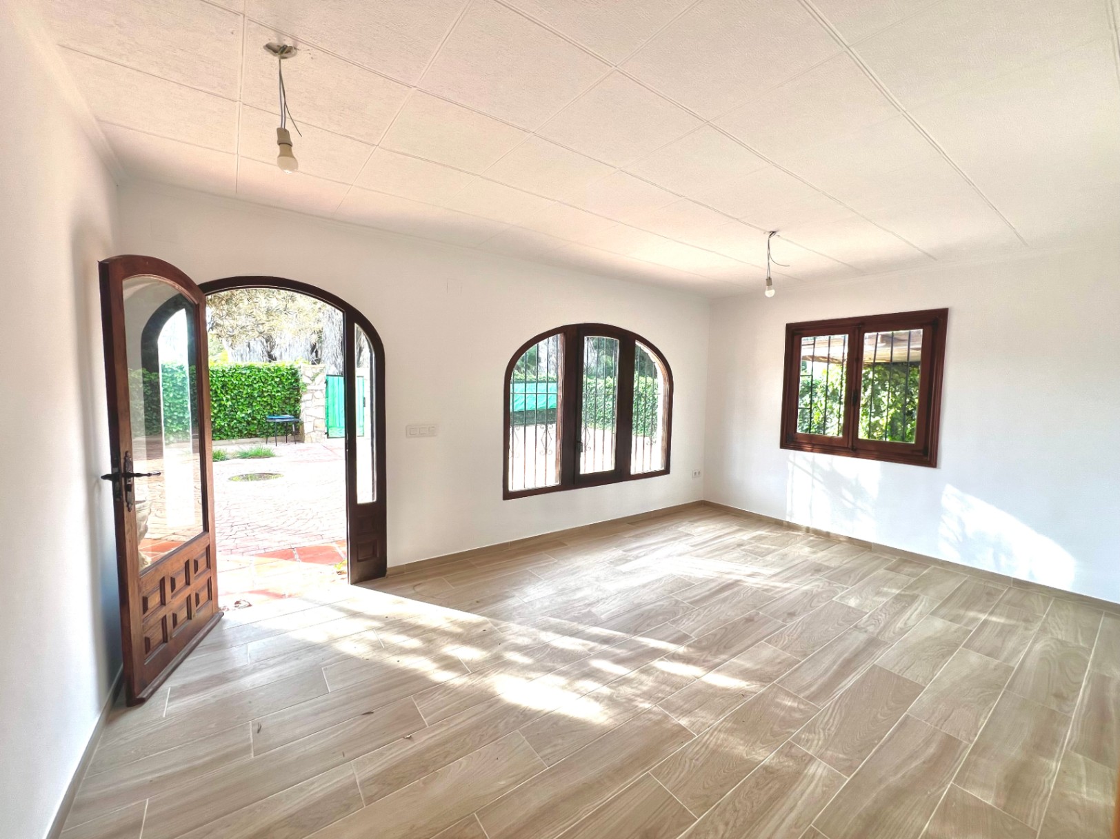 Bright living area featuring wood-effect floor tiles, arched wooden window frames with security bars, and a matching arched door opening onto the paved garden patio.