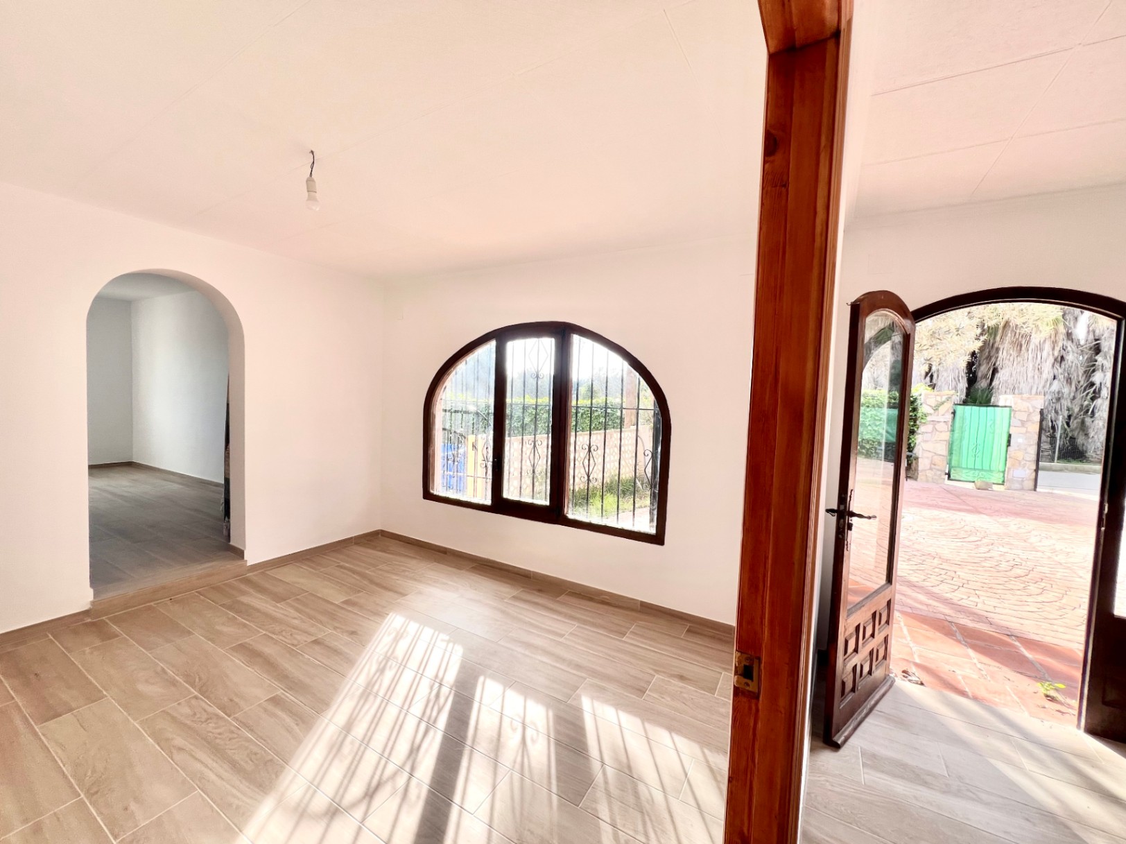 Bright living space featuring wood-effect tile flooring, a classic arched window, and a wooden door opening onto the paved outdoor patio.