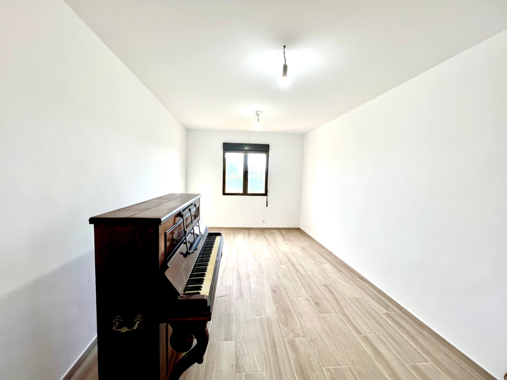 Elongated room featuring wood-effect tile flooring, white walls, and a vintage upright piano. A dark-framed window provides natural light at the far end.