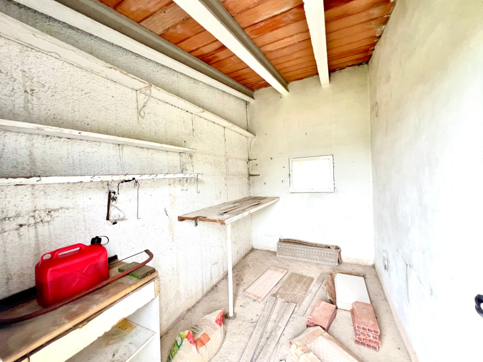 Functional storage room featuring exposed wooden beams and terracotta ceiling tiles. Equipped with wall shelving, a workbench, and a small window for natural light.