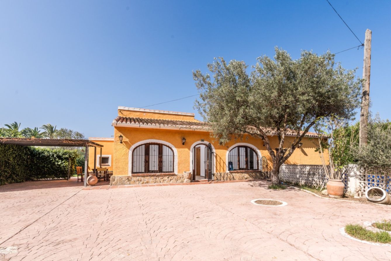 Mediterranean-style facade featuring ochre walls, arched windows, and stone detailing, set against a spacious paved patio with a mature olive tree.