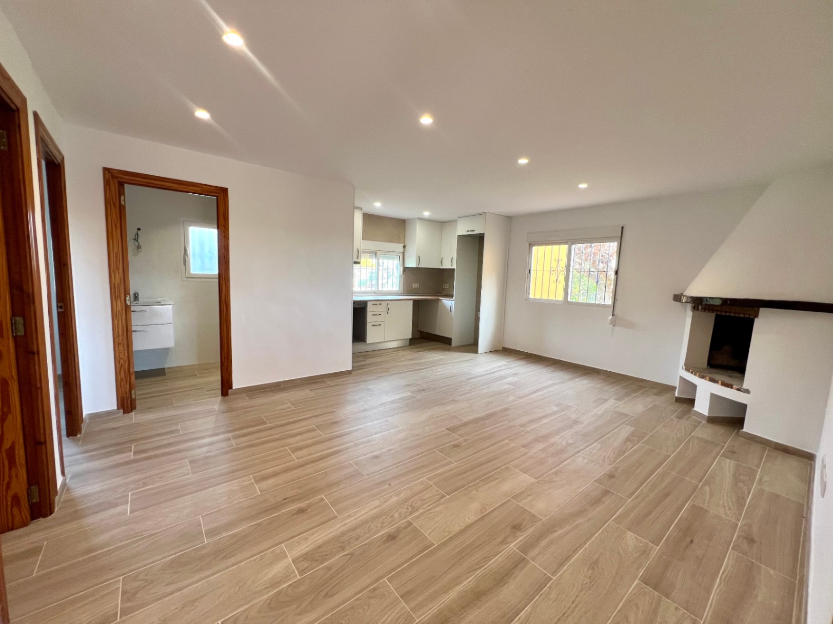 Open-plan living area featuring a corner fireplace, integrated white kitchen, and wood-effect ceramic flooring with recessed lighting.