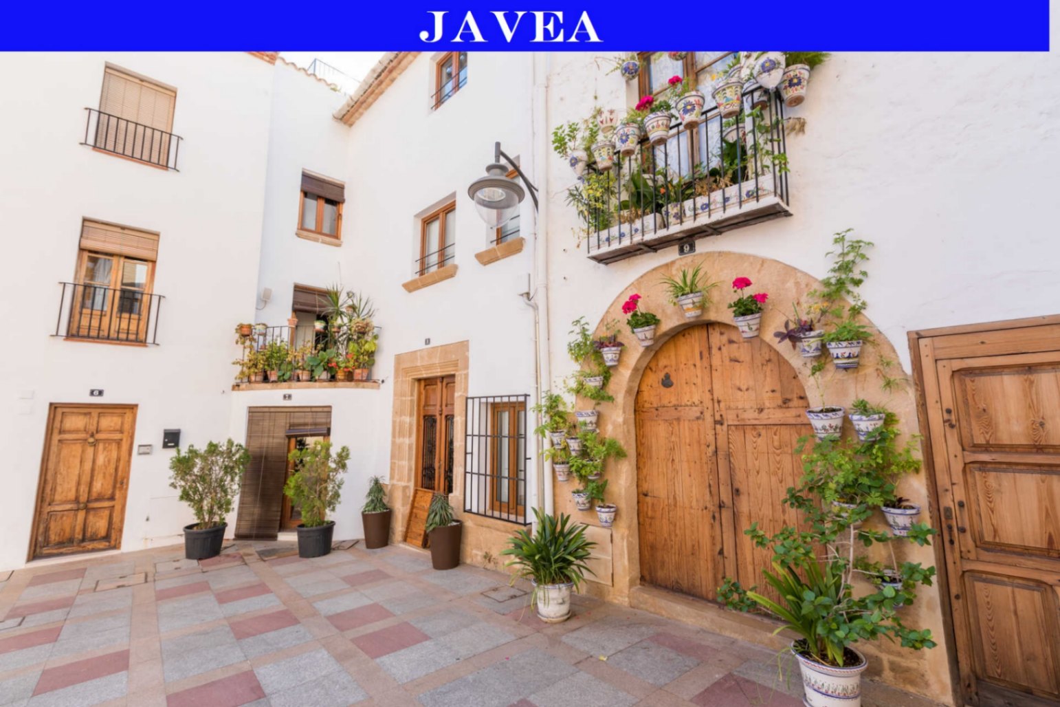 Traditional Javea facade featuring a large arched wooden door with stone framing, adorned with ceramic flower pots and wrought iron balconies.