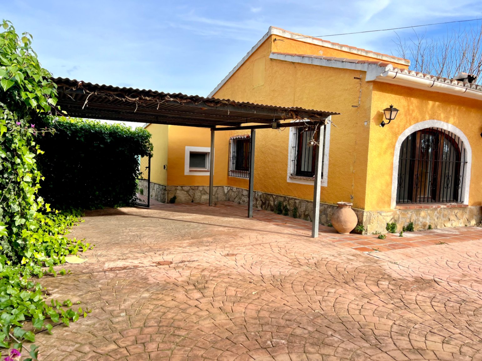 Yellow facade featuring a natural stone base, a covered carport, and fan-patterned paving adjacent to an arched window with wrought iron bars.