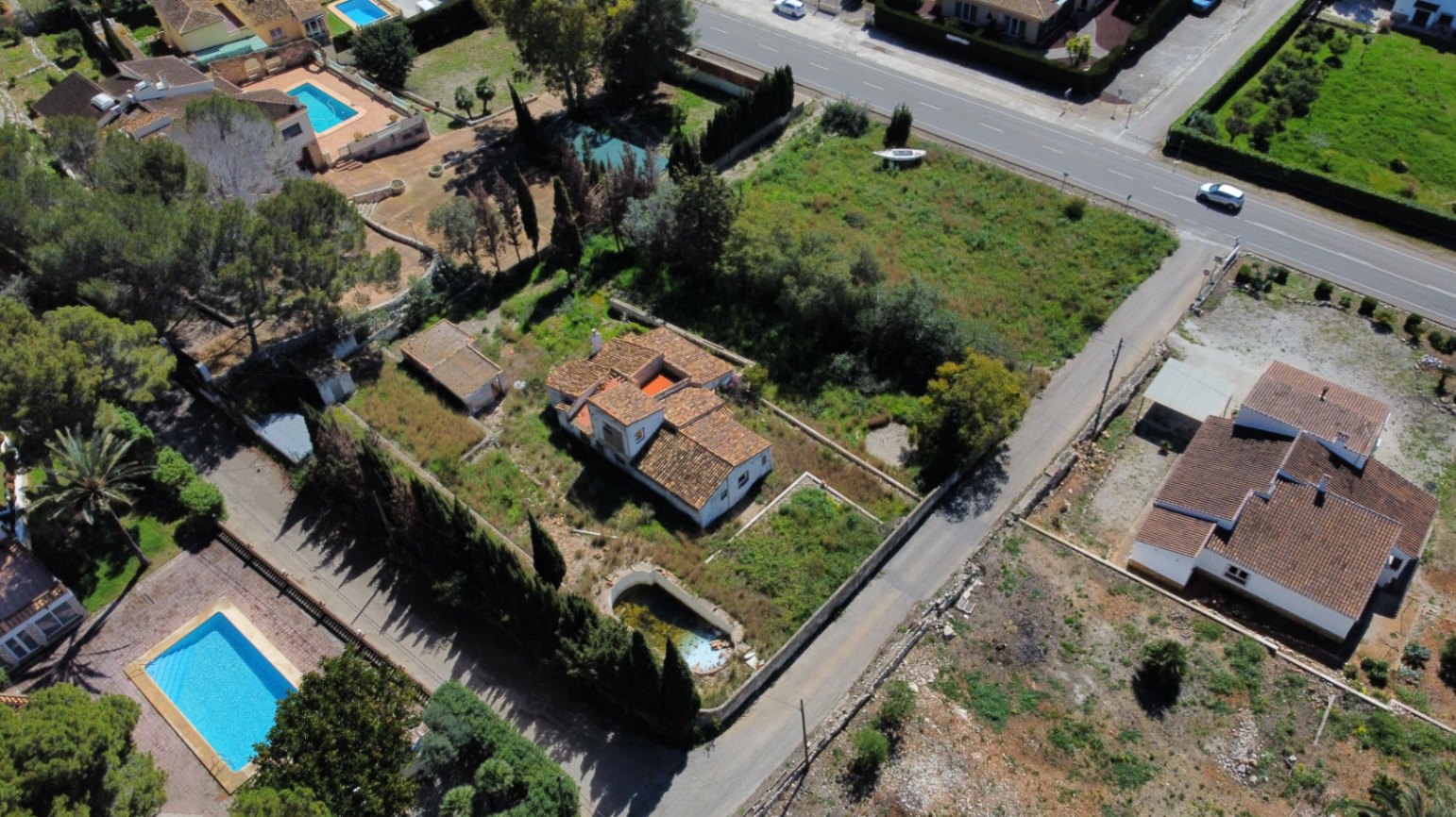 Aerial perspective of the 2,895sqm plot featuring the traditional villa, a pool in need of repair, and expansive grounds with Mediterranean trees and road access.