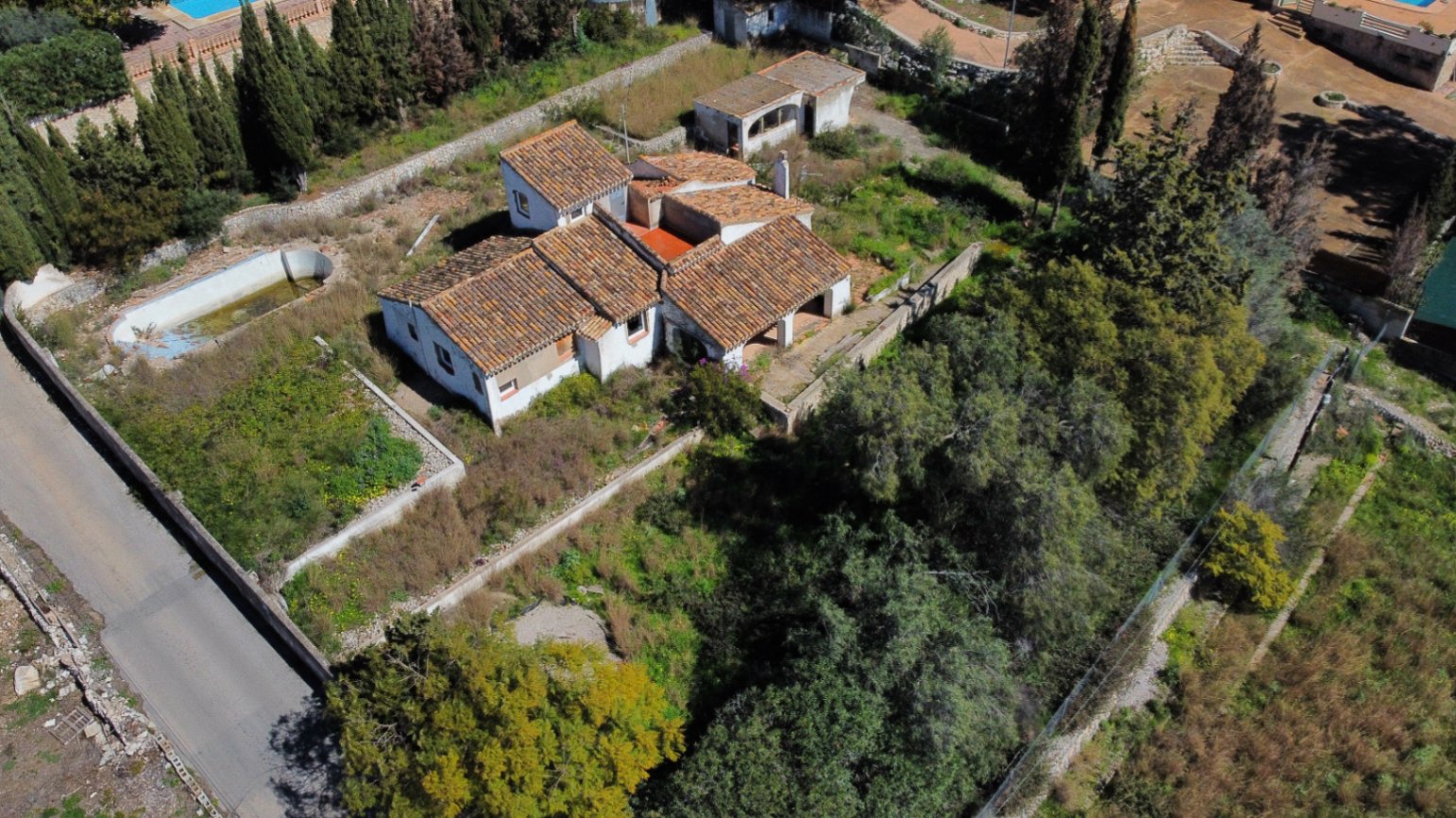 Aerial view showing the multi-level terracotta roof structure, the expansive plot with mature trees, and the existing pool area requiring renovation.