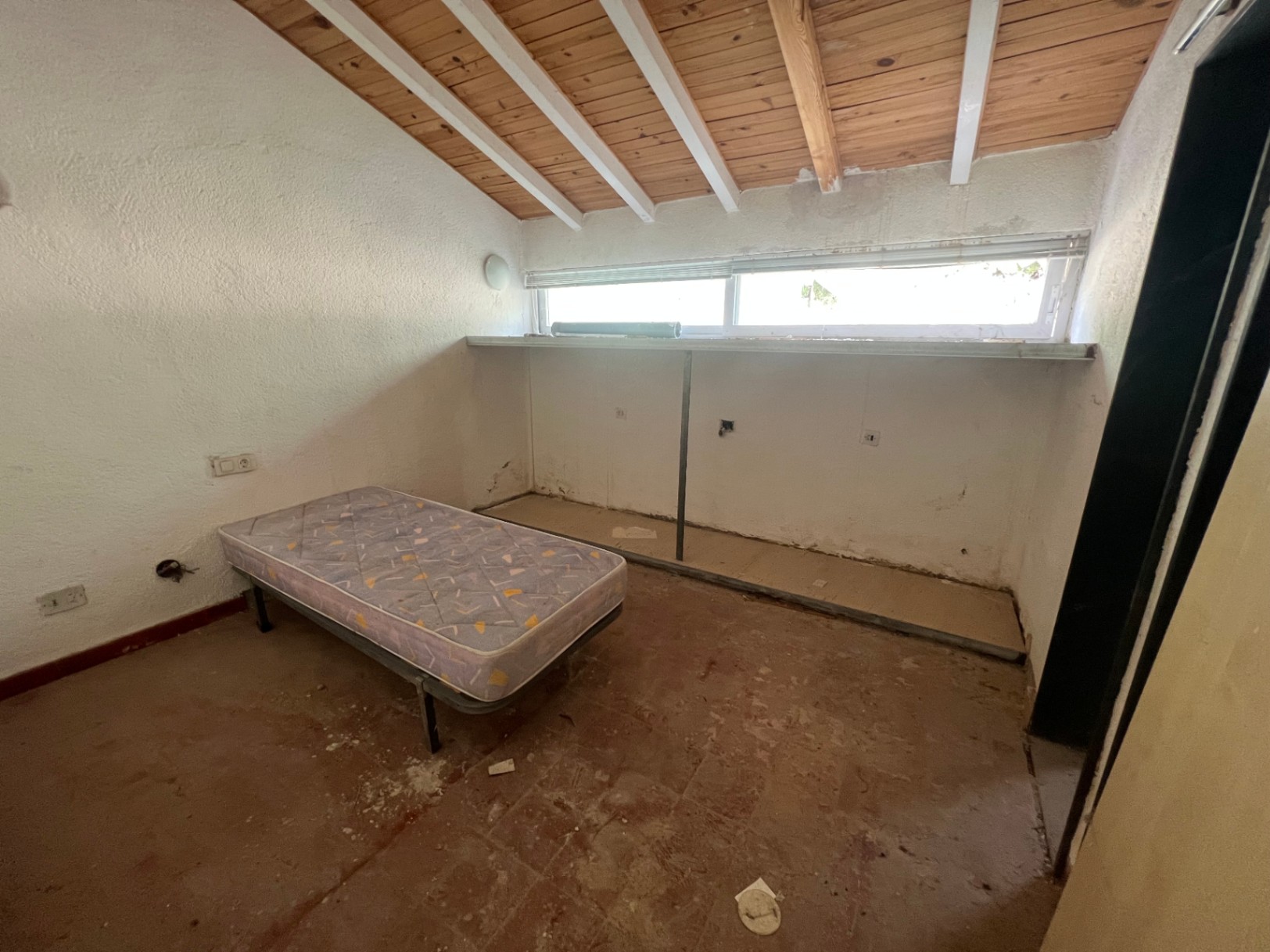 Bedroom featuring a sloped ceiling with exposed wooden beams and white rafters. The space includes original clay tile flooring and a long horizontal window.