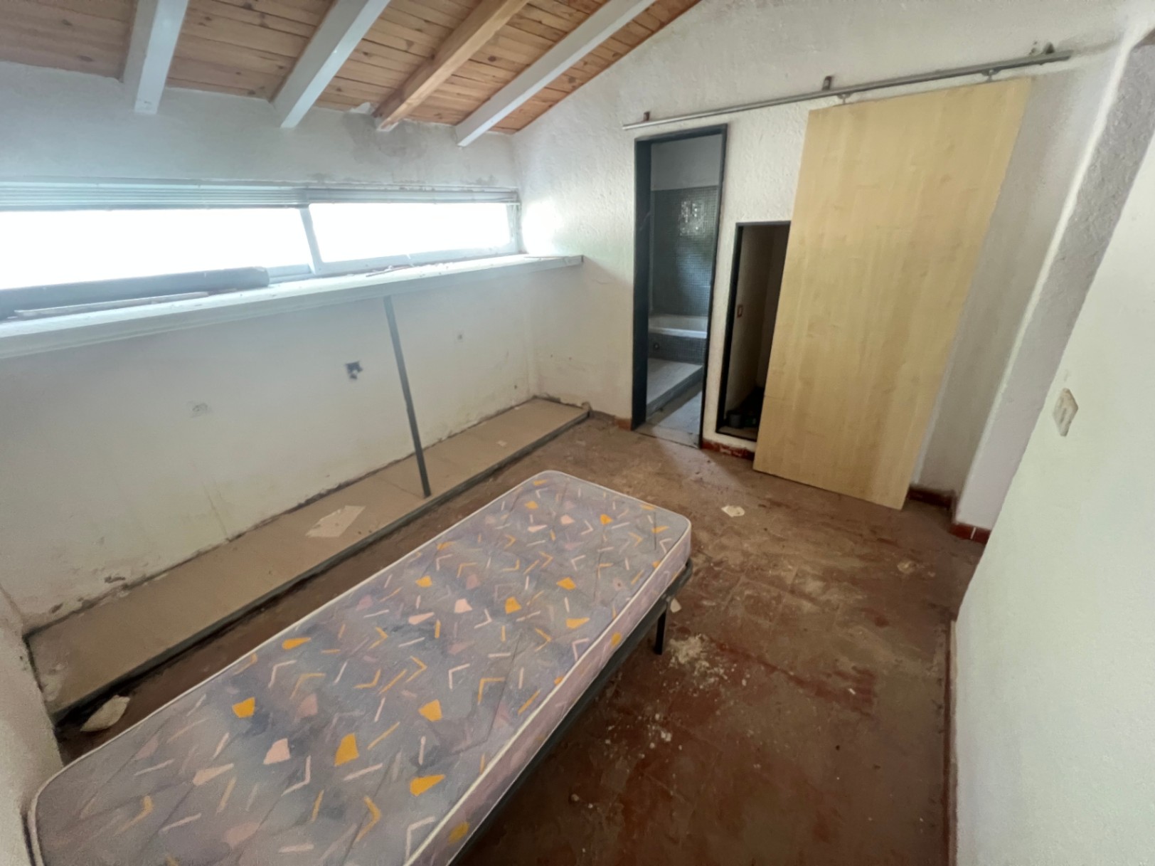 Bedroom featuring exposed wooden ceiling beams, a long horizontal window, and an ensuite bathroom. The space offers potential for a complete renovation.