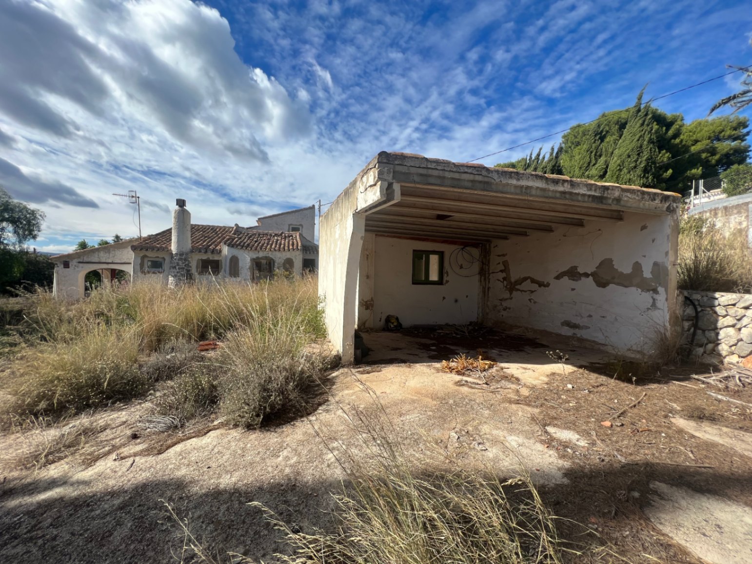 Detached carport and storage structure requiring renovation, set within a large plot with dry Mediterranean vegetation and the main villa visible in the background.