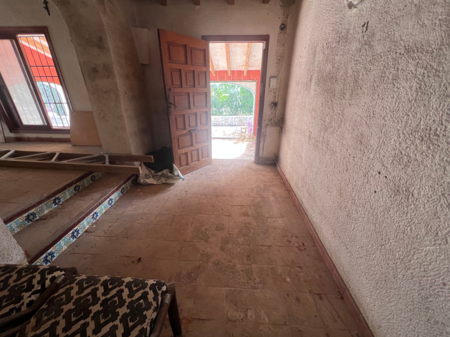 Entrance hall featuring terracotta flooring, a paneled wooden door, and stairs with decorative ceramic risers. A bright space ready for a complete renovation.