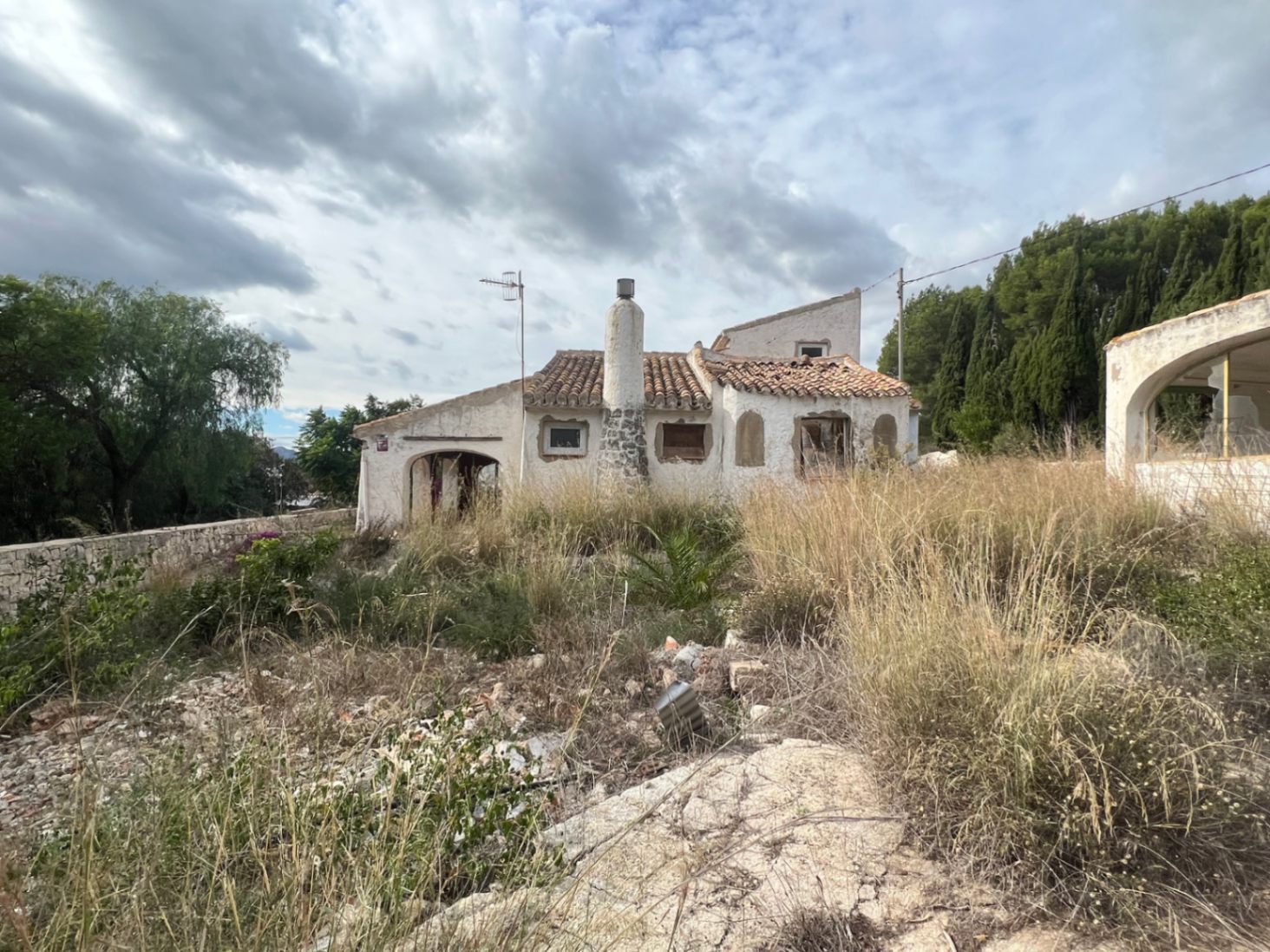 Exterior view of a rustic villa in Jávea featuring a stone-based chimney, terracotta roof, and traditional arches on an overgrown plot awaiting renovation.