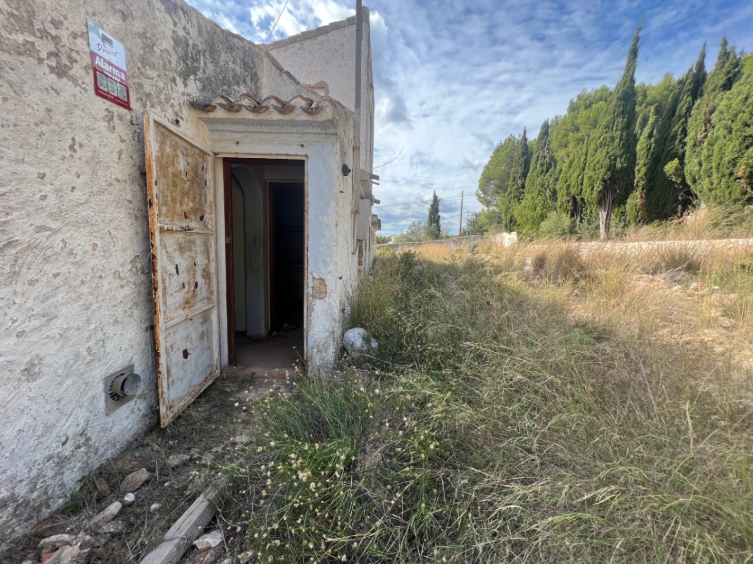 Exterior view of a secondary structure featuring a weathered metal door and original masonry, set within an expansive plot with wild Mediterranean vegetation.