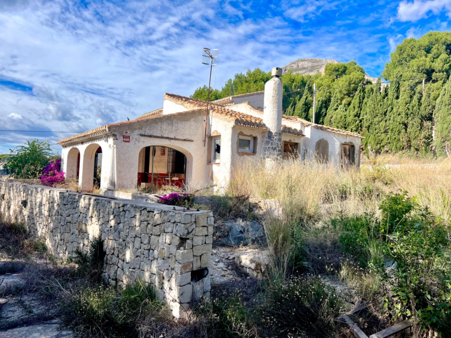 Exterior view of the villa featuring an arched naya, natural stone wall, and terracotta roof, set against the backdrop of the Montgó mountain.