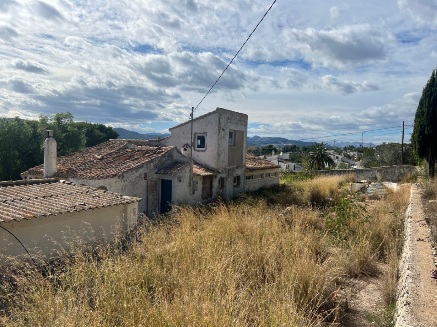 Exterior view of the villa featuring traditional ceramic tile roofs and a weathered facade, set on a large plot with natural vegetation and distant mountain views.