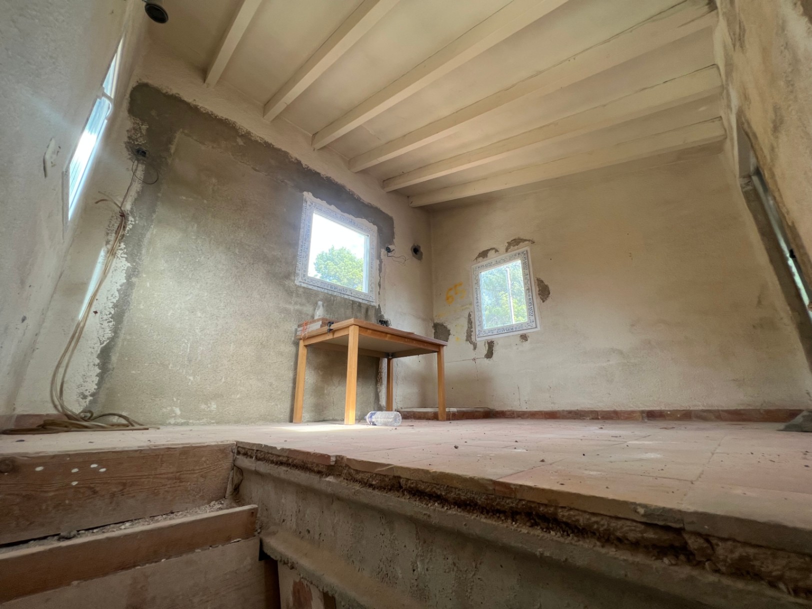 Interior room under renovation featuring exposed ceiling beams, traditional terracotta flooring, and unfinished walls. Includes new window frames and exposed wiring.