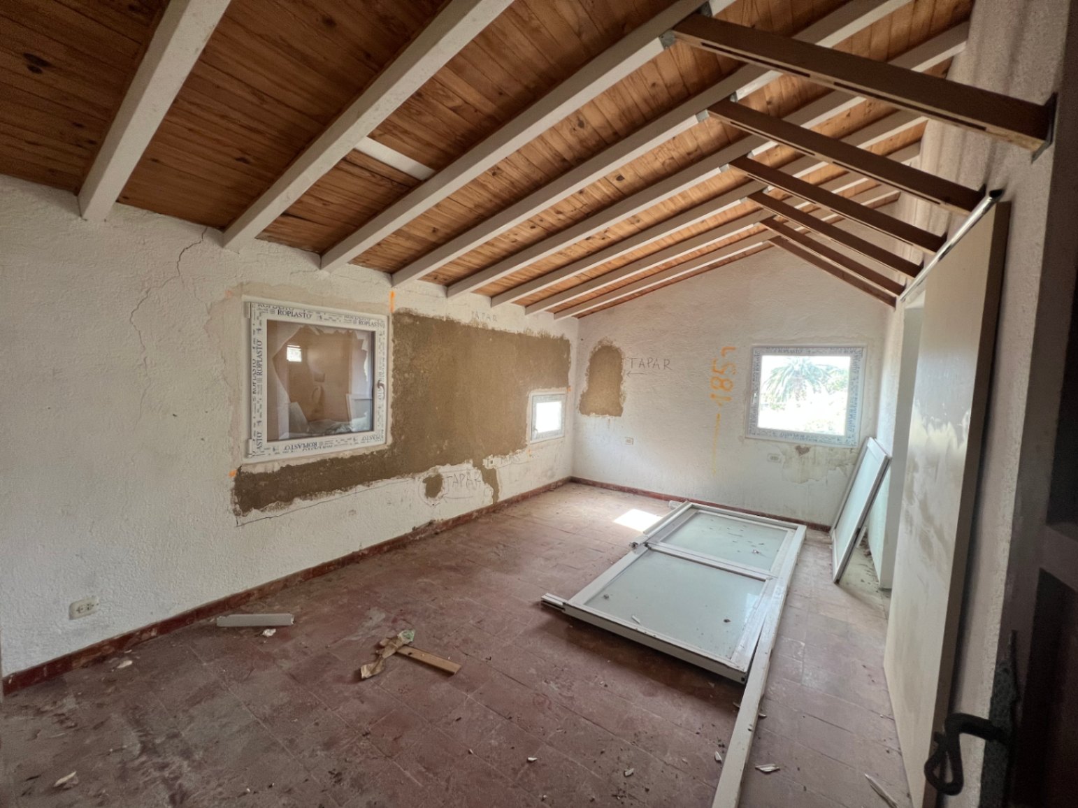 Interior room undergoing renovation featuring sloped wooden ceilings with white beams, terracotta tiling, and new PVC window frames being installed.
