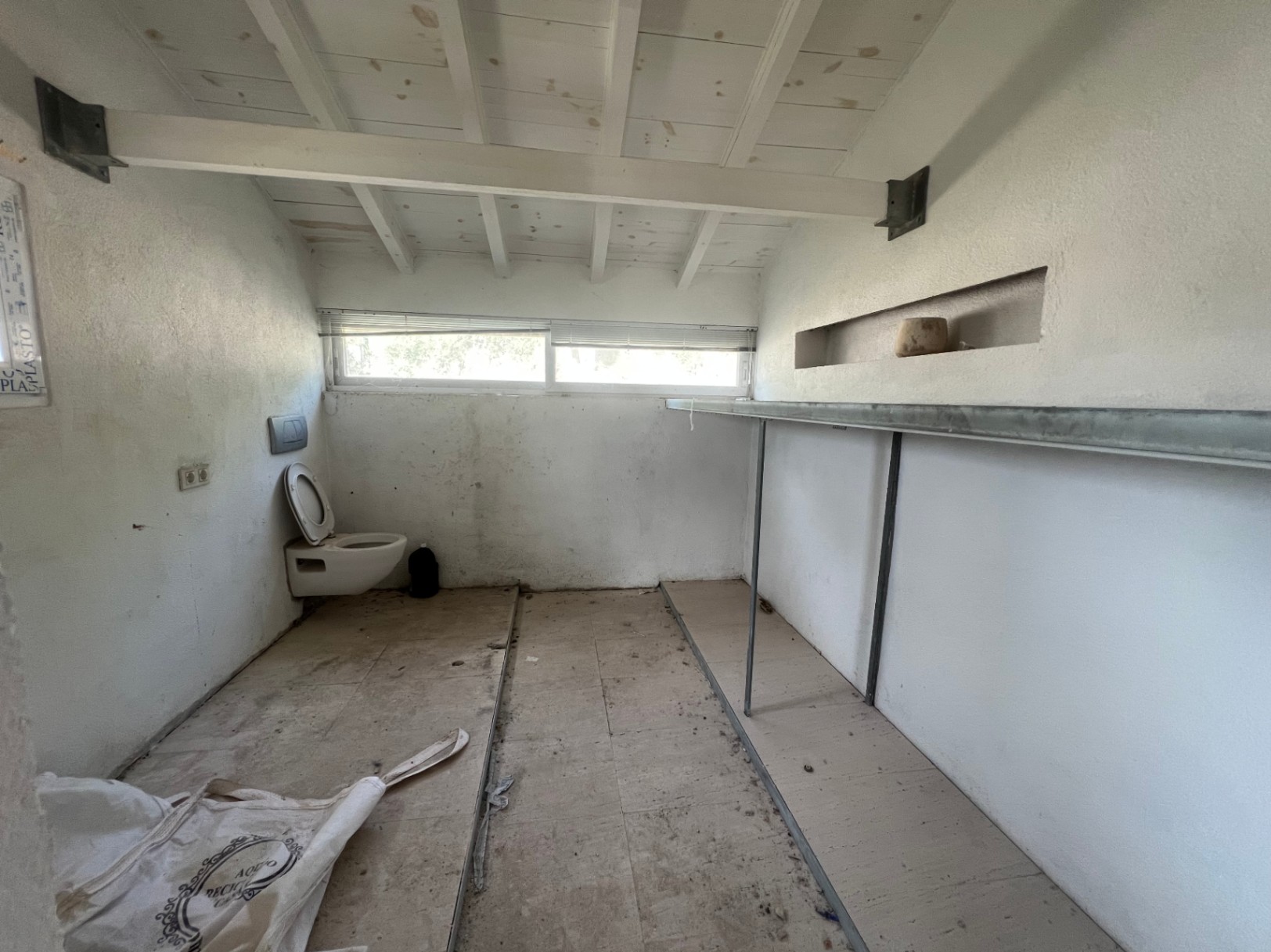 Interior space for renovation featuring white wooden ceiling beams, a wall-mounted toilet, and fixed shelving. A horizontal window provides natural light.