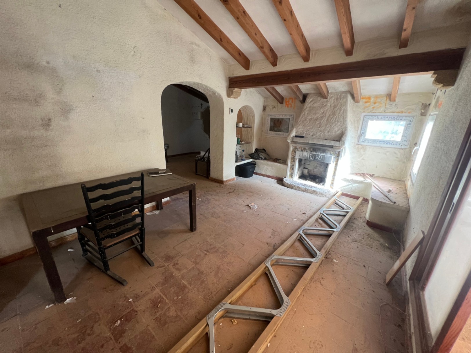 Rustic living area awaiting renovation, featuring exposed wooden beams, a traditional fireplace, and terracotta floors. Arched doorways lead to adjacent rooms.