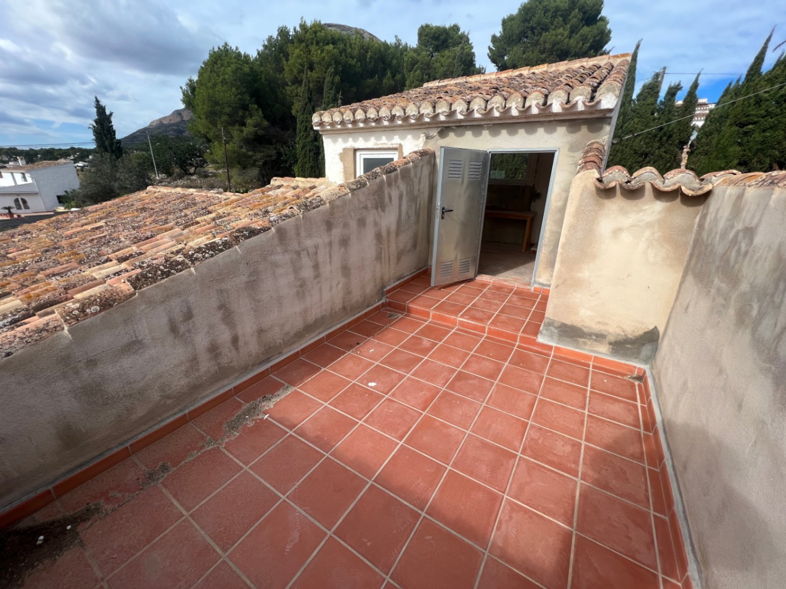 Upper terrace featuring terracotta tiling and access to a small independent outbuilding with a pitched clay tile roof, surrounded by Mediterranean vegetation.